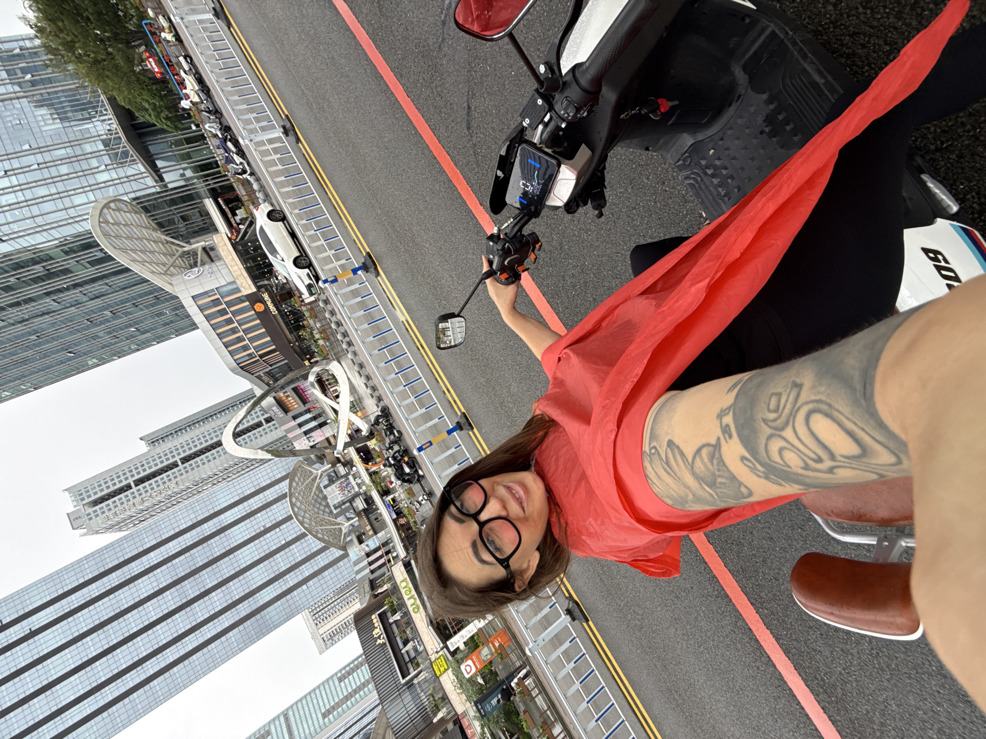 A woman with dark hair, large black-rimmed glasses, and a red shawl over her shoulders is sitting on a white scooter. She is looking at the camera and smiling. She has tattoos on her left arm. The scooter's digital display shows a speed of 0. The background shows a city street with tall buildings, shops, and other scooters parked along the sidewalk. The street is wet, indicating it may have recently rained. A white car is visible in the distance. The overall scene appears to be a street in Huizhou, China, likely a commercial area given the presence of shops and modern architecture.
