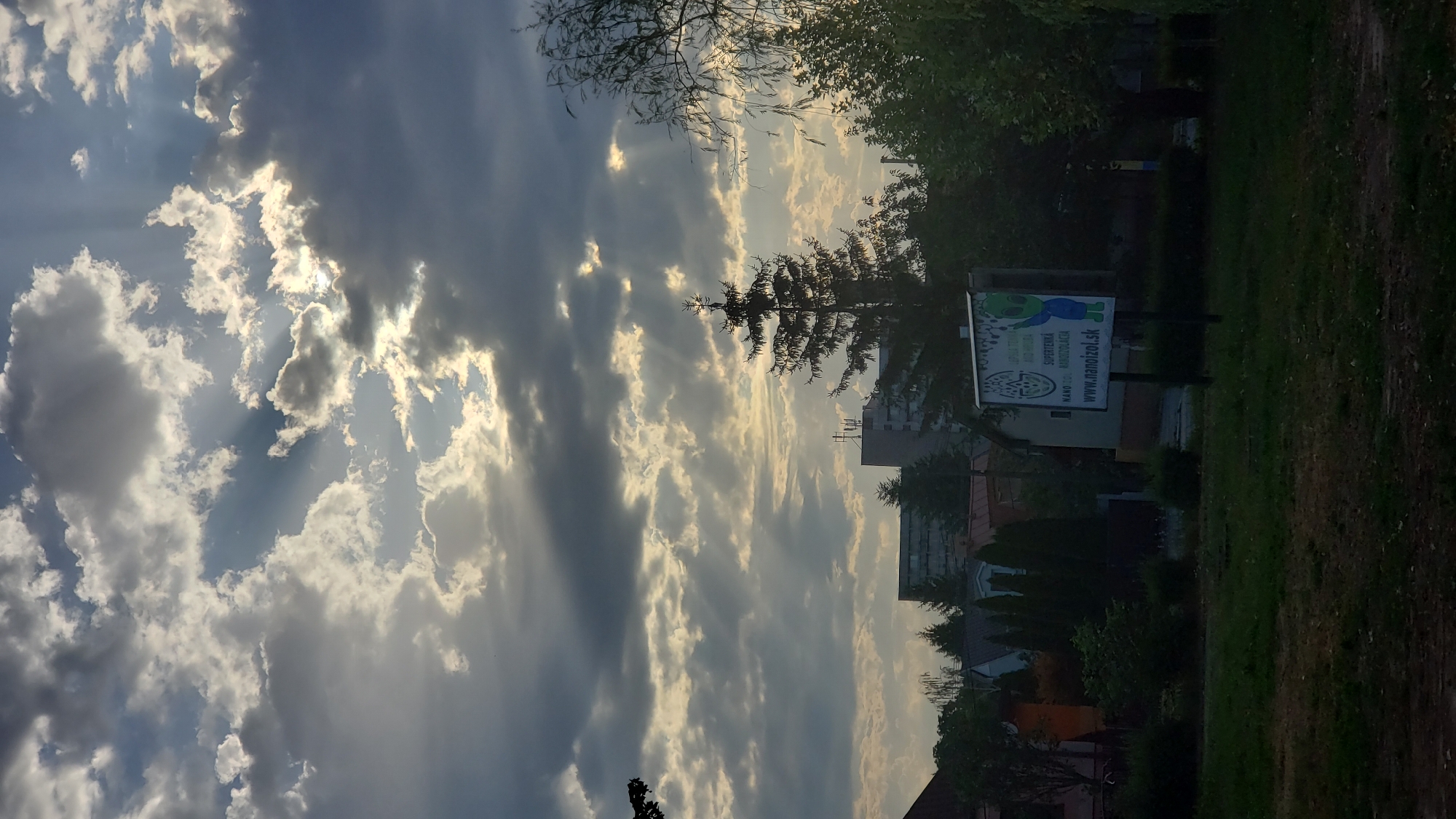 The image captures a view of a cloudy sky with sunbeams breaking through, over a suburban area. In the foreground, a grassy area is visible, with some trees and bushes. In the middle ground, there are several residential buildings and a tall evergreen tree. To the right, a billboard with an alien-like character and text, including "SUPERTEHNIKA" and "www.nanoizol.sk", is prominently displayed. The background shows a larger building with antennae, characteristic of a European city. The lighting suggests it is either early morning or late afternoon. The location appears to be a residential district in Prešov, Slovakia, likely near the sídlisko Sekčov area, given the architectural style of the buildings and the presence of such billboards.