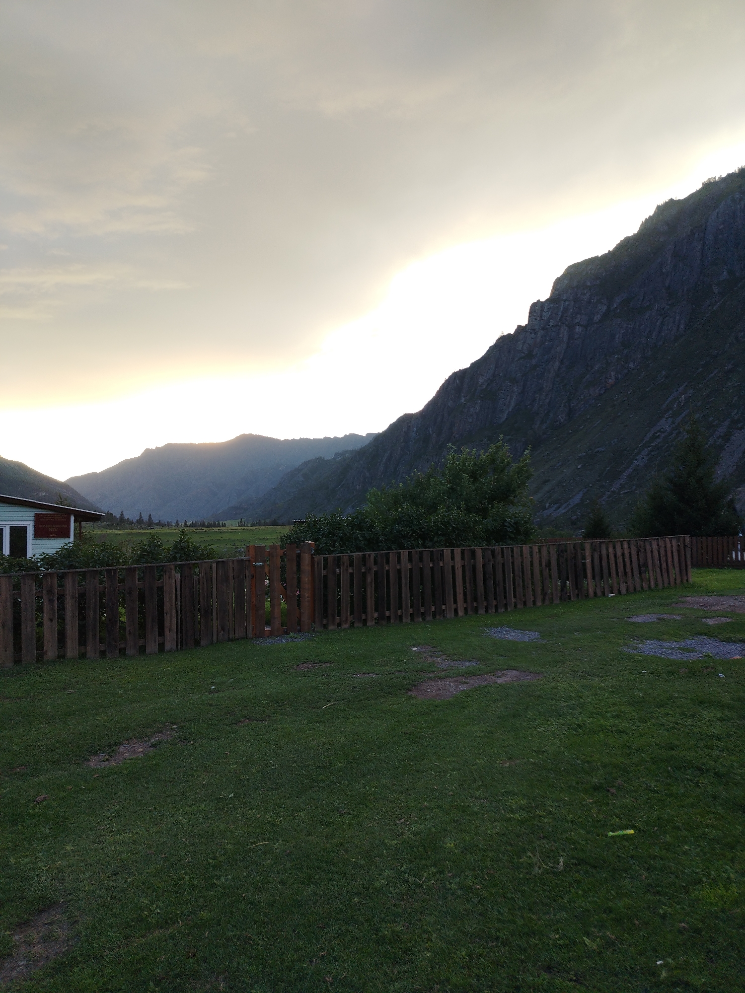 The image depicts a scenic outdoor scene, likely in Iodro, Russia, at dusk or dawn. A wooden fence runs horizontally across the mid-ground, separating a grassy area in the foreground from a more vegetated area beyond. To the left, a light-colored building with green trim is partially visible. In the background, rolling hills and a steep, rocky mountain range dominate the landscape. The sky is overcast with a soft glow indicating the sun is near the horizon. The overall impression is one of tranquility and natural beauty.