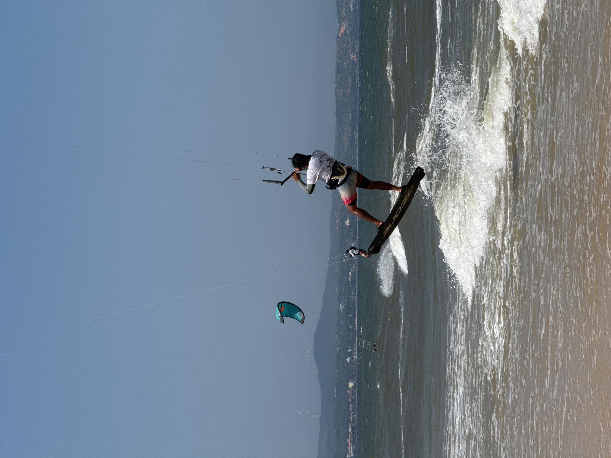 An action shot captures a kitesurfer mid-air, performing a jump over choppy ocean waves. The kitesurfer, positioned on the right side of the frame, is wearing a white t-shirt, colorful shorts, and a harness. They are holding onto the control bar, which is attached to a kite that is out of frame. Their board is angled upwards, with spray erupting from the wave below.
In the background, a second, smaller kite is visible in the upper left quadrant, appearing as a teal and orange diamond shape. Further in the distance, a hazy coastline with rolling hills is dotted with buildings and wind turbines. The sky is a clear, pale blue, suggesting a sunny day. The foreground is dominated by the sandy beach, blurred by the action of the waves. The image is set in Mũi Né, Vietnam, known for its kitesurfing and windsurfing conditions.