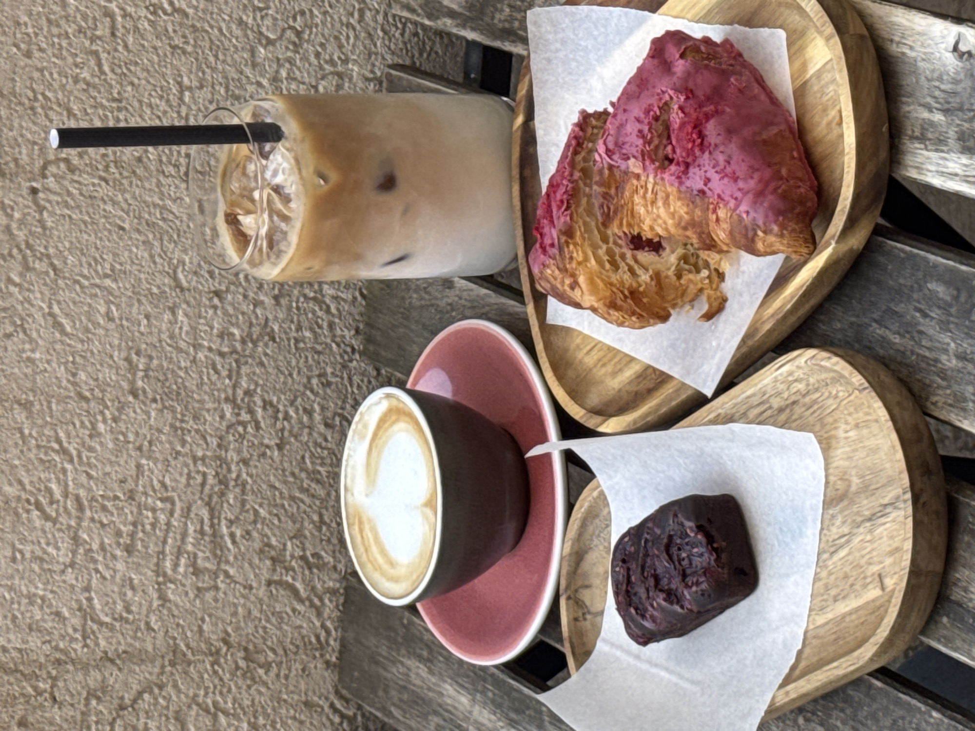 This overhead shot captures a delightful outdoor cafe scene in what appears to be Budva, Montenegro. The subjects are an assortment of beverages and pastries neatly arranged on a wooden slatted table or bench.
On the left, a tall glass holds iced coffee with ice cubes and a black straw. To its right, an open-faced croissant, generously topped with a vibrant pink glaze or icing, rests on a small wooden serving plate lined with white parchment paper. Below the iced coffee, a dark brown cup containing a hot coffee, likely a latte or cappuccino given the visible latte art heart design, sits on a pink ceramic saucer. Next to this, on another small wooden plate also lined with white parchment paper, is a dark, dense pastry, possibly a chocolate brownie or a berry-infused treat, showcasing a rich, textured surface.
The activity depicted is a casual coffee break or snack time, suggesting a moment of relaxation and enjoyment. The natural lighting indicates it is daytime. The background features a light-colored, textured surface, possibly a stucco wall or rough concrete, contributing to an inviting outdoor ambiance. There is no visible text in the image. The overall impression is one of a pleasant and aesthetically arranged culinary experience.