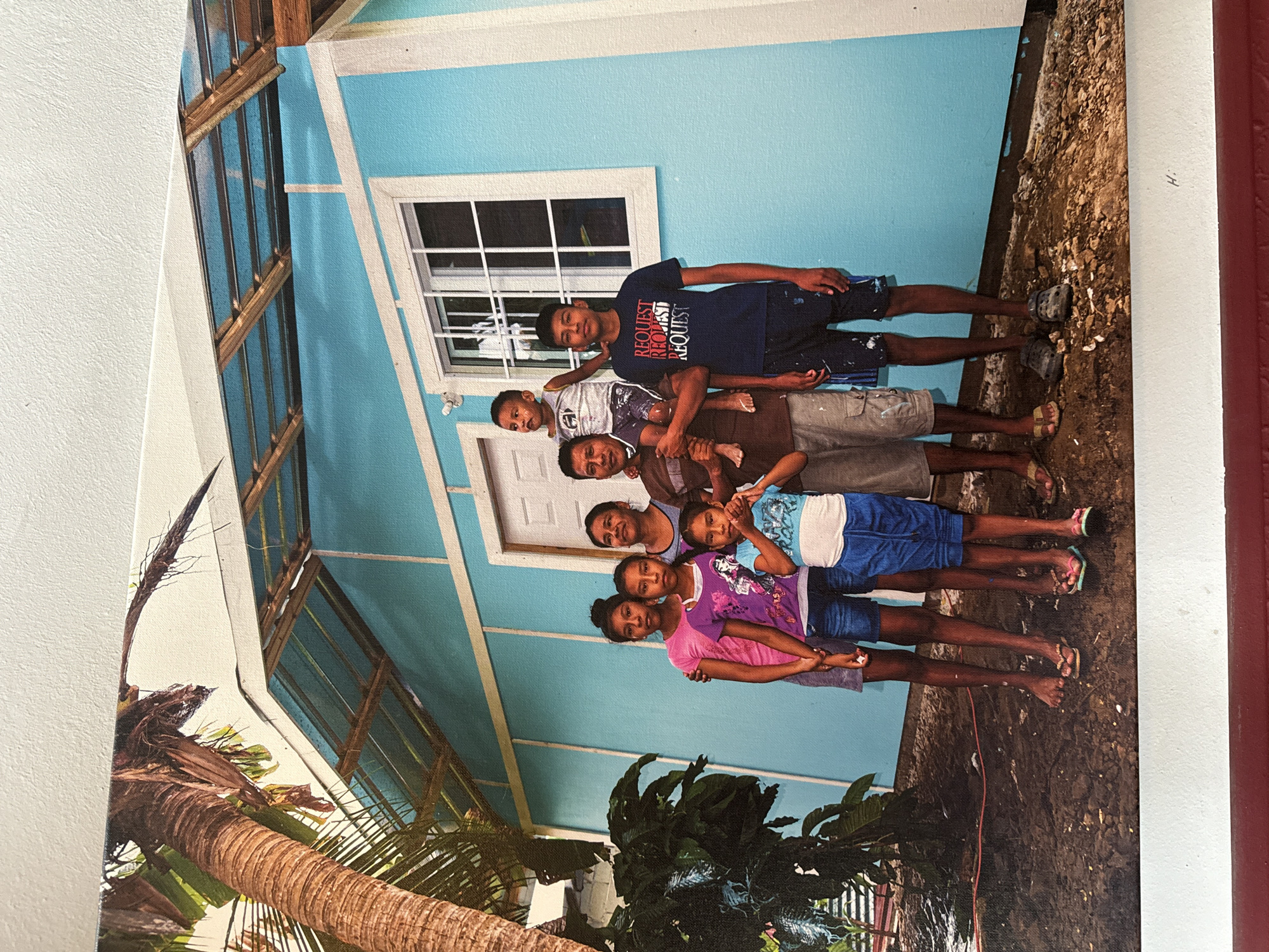 In an outdoor setting, a family of six poses in front of a newly constructed, bright blue wooden house. The house has a white trim around a window and a white door. A palm tree is visible to the left of the house.
The family consists of three girls, two boys, and one adult male and one adult female. The girls are in the front, with one in a pink shirt and shorts, another in a purple t-shirt and denim shorts, and the smallest in a blue and white t-shirt and blue shorts. Behind them are two adult women, one in a patterned shirt, and the other in a brown shirt. To the right is a boy in a dark blue t-shirt with red lettering and dark shorts. The man is on the far right, wearing a brown shirt and khaki shorts, and holding a young boy on his shoulders. The young boy is wearing a white t-shirt and shorts. The ground in front of the house is dirt with some scattered debris.
The image does not provide any specific information about the city of Bad Blankenburg, Germany.