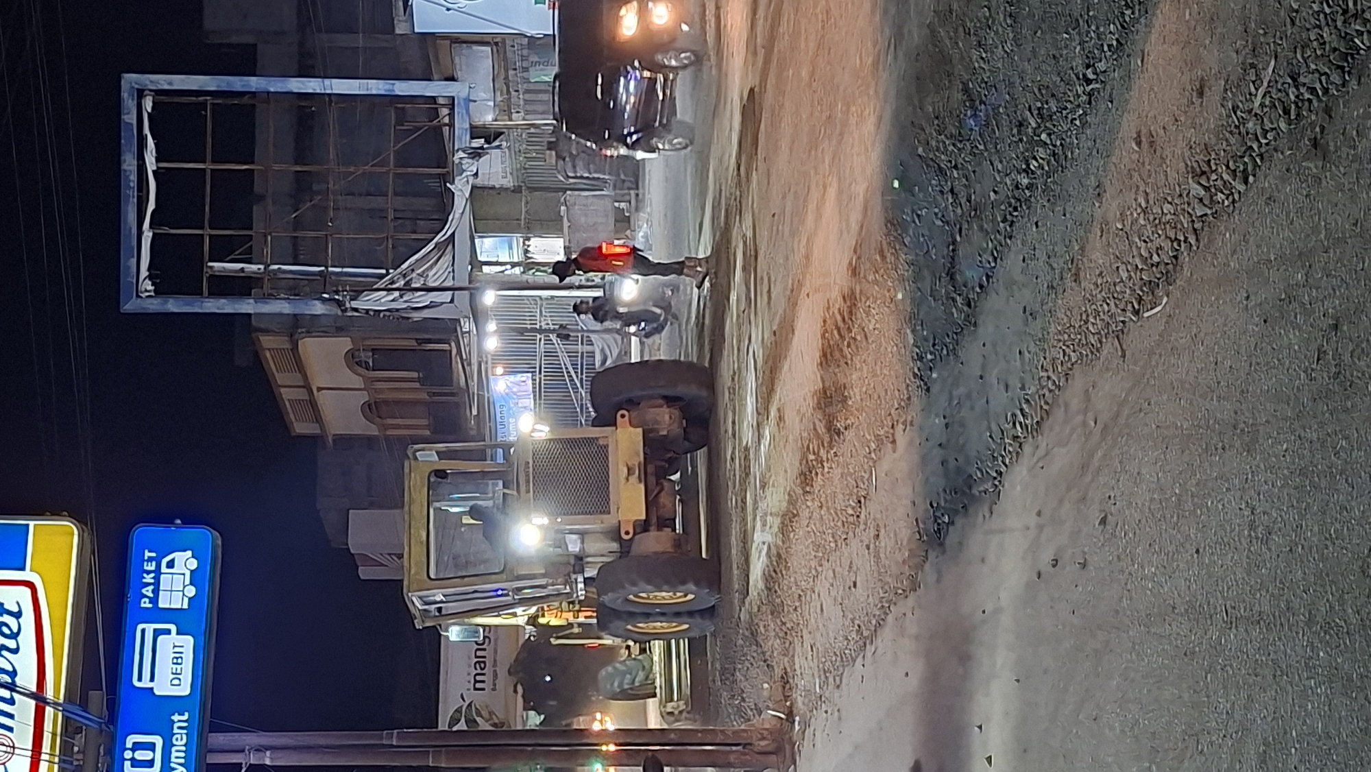 A nighttime scene in Blangpeuria, Indonesia, depicts a street with several vehicles and people. The image is vertically oriented. In the foreground, a yellow backhoe loader is parked, its large tires prominent. To the left, a blue and yellow sign for "Indomaret" is visible, featuring the text "PAKET" and "DEBIT" along with icons for a delivery truck and credit cards. Further back, a dark-colored car with illuminated headlights is present. Several individuals are scattered throughout the scene. One person, wearing a red shirt, stands near the car. Another person is seen riding a motorcycle. The environment appears to be a street with a rough, gravelly surface. The lighting suggests it is nighttime, with artificial lights illuminating the area. The overall atmosphere is that of a typical street scene in a semi-urban or rural Indonesian setting at night.