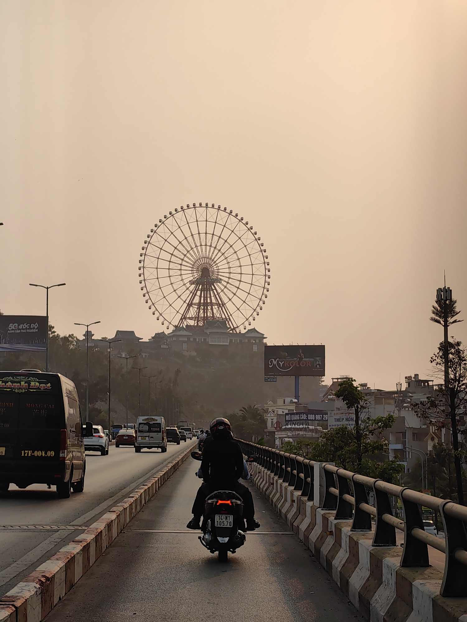 The image captures a street scene in Hong Gai Ward, Vietnam, distinguished by a large Ferris wheel in the background. The sky is a hazy yellow, suggesting either sunset or a smoggy atmosphere.
A motorbike with two occupants is in the foreground, moving along the road. It has a license plate visible as "14-M1 105.70". A van with license plate "17F-004.09" is traveling in the same direction, slightly ahead of the motorbike. Several other vehicles are further ahead on the road.
Streetlights and advertisements are present on the roadside. Advertisements for "Mykolor" and "5G Tốc Độ" are visible. The Ferris wheel stands atop a hill with buildings constructed around it. Trees and other greenery are scattered throughout the urban landscape. A cell tower stands near the right edge of the frame. The road is divided by a median barrier with red and white blocks and a metal railing.
