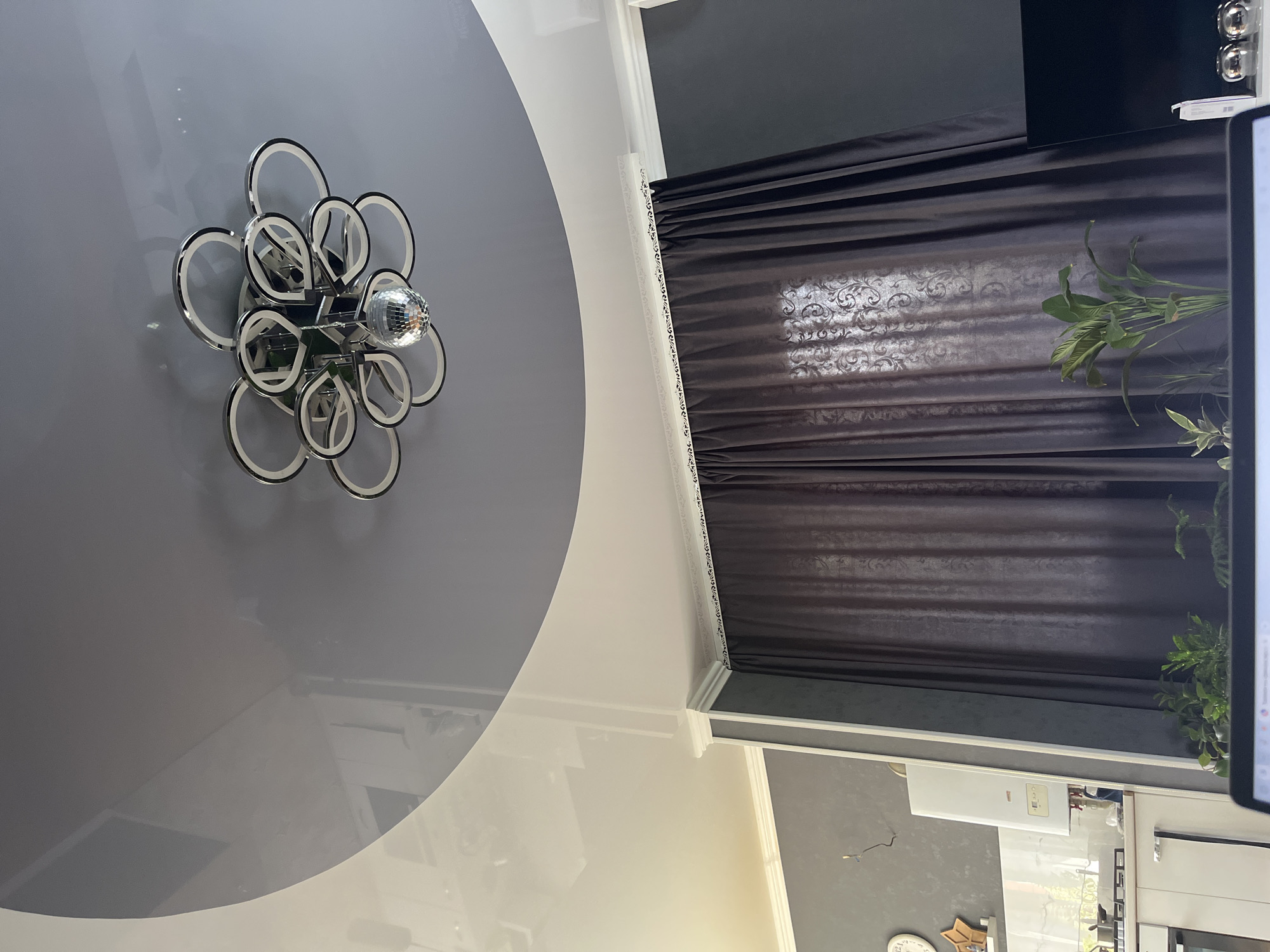 The image captures an interior view, focusing on a modern ceiling with a distinctive dual-toned design, featuring a glossy white section and a matte grey section. A contemporary chandelier with multiple ring-shaped lights and a central disco ball hangs from the grey section. Below the ceiling, dark brown, patterned curtains are drawn across a window, obscuring the view outside. To the left, a glimpse of a kitchen area is visible, showing white cabinets, a white gas water heater, and a round clock on the wall. Several potted plants are placed in front of the curtains. A portion of a laptop screen is visible in the foreground, suggesting the photograph was taken from the perspective of someone using a laptop. There is no visible indication of the city of Saratov, Russia, nor are there any people or specific actions depicted beyond the static arrangement of objects.