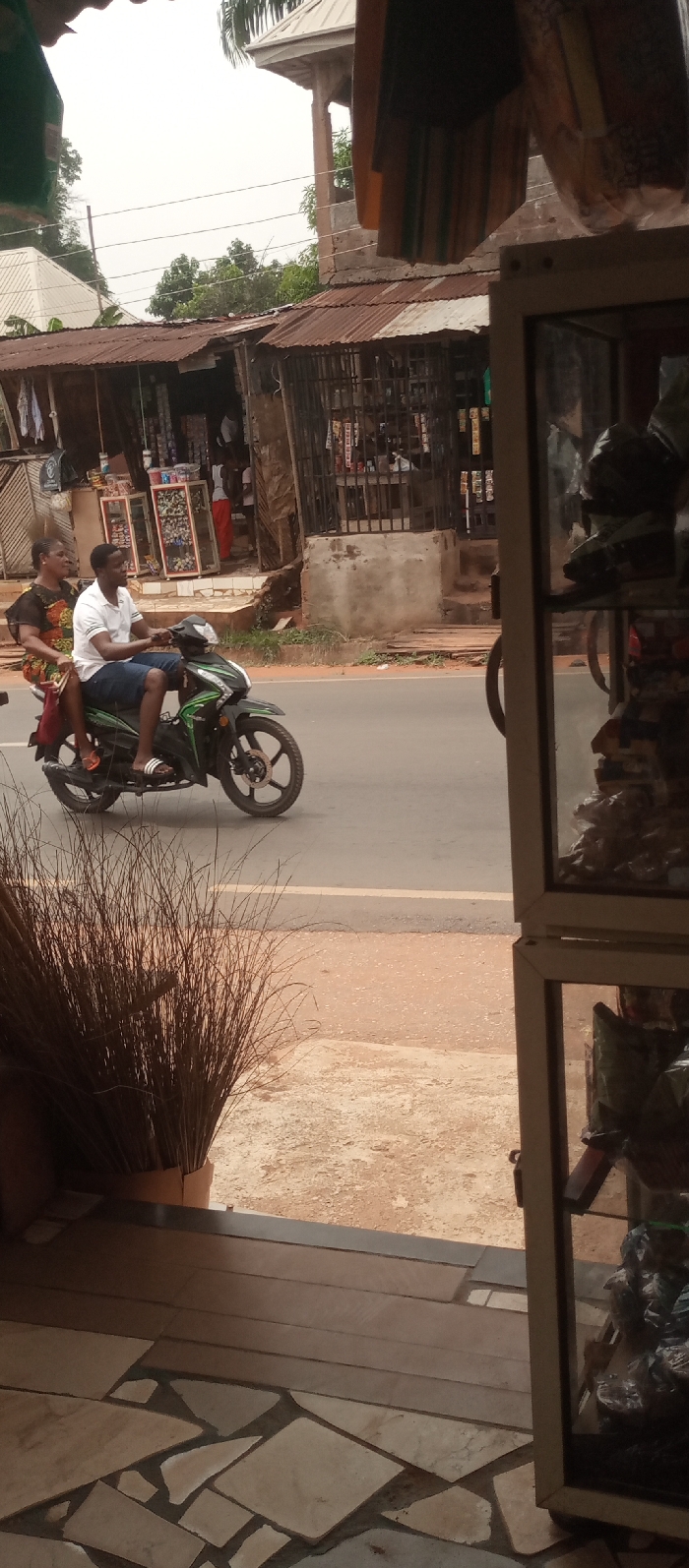 The image portrays a street scene in Umuoji, Nigeria, viewed from what appears to be an indoor space. In the immediate foreground on the left, a bundle of tall, dark, dried plant stalks stands. To the right, a section of a light-framed display case, filled with various objects, is visible. The interior floor features an irregular mosaic pattern of light-colored tiles.
The midground is dominated by a paved asphalt road, marked with a faint white line. A dark green and black motorcycle is in motion, traveling from right to left across the frame. The motorcycle is operated by a male driver wearing a white polo shirt, blue shorts, and white-striped slides. A female passenger, dressed in a colorful patterned outfit and orange sandals, is seated behind him, holding a red bag. Both occupants are facing forward.
The background reveals the opposite side of the street, populated by several structures. These include low-rise buildings or market stalls, predominantly featuring rusted corrugated metal roofs. One establishment on the left displays clothing items externally. A central shop or stall exhibits various packaged goods, visible behind a metal grate. Further back and partially obscured, a taller concrete structure with a light-colored, pointed roof rises above the lower buildings. Green trees and foliage are interspersed throughout the background under a light, overcast sky.
