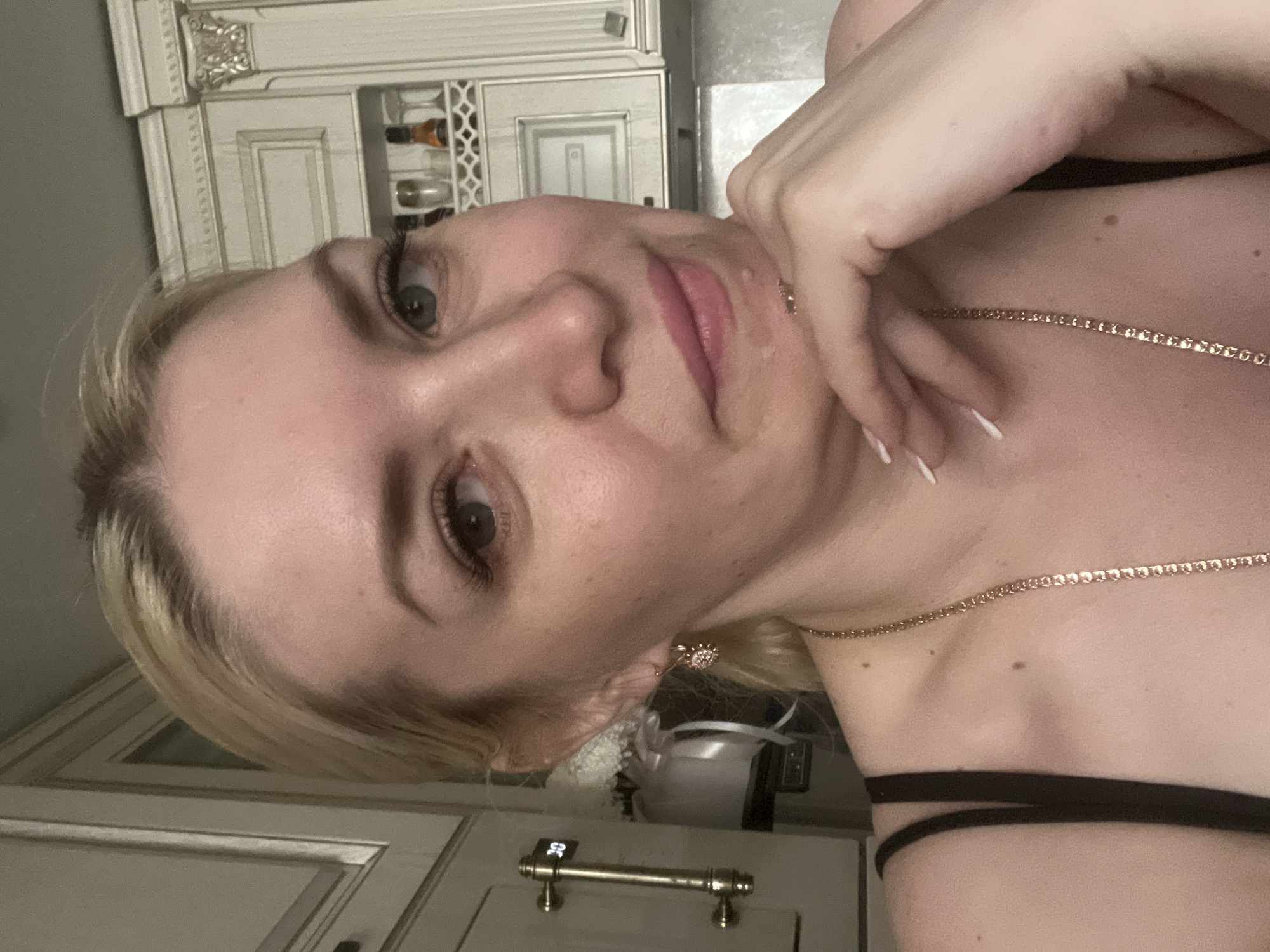 This is a close-up, shoulder-up photograph of a light-skinned woman with blonde hair, likely in her 30s, posing for a selfie. She is wearing a black camisole and a gold chain necklace. Her left hand is resting on her chin, and her fingernails are painted white. She has light blue eyes and is wearing subtle makeup. The background appears to be a kitchen with cream-colored cabinets and decorative molding. On the left side of the image, a metallic handle with a digital display showing "30" is visible on a cabinet. The overall lighting is warm and indoor. The image does not provide any specific clues about the location within Tyumen, Russia.