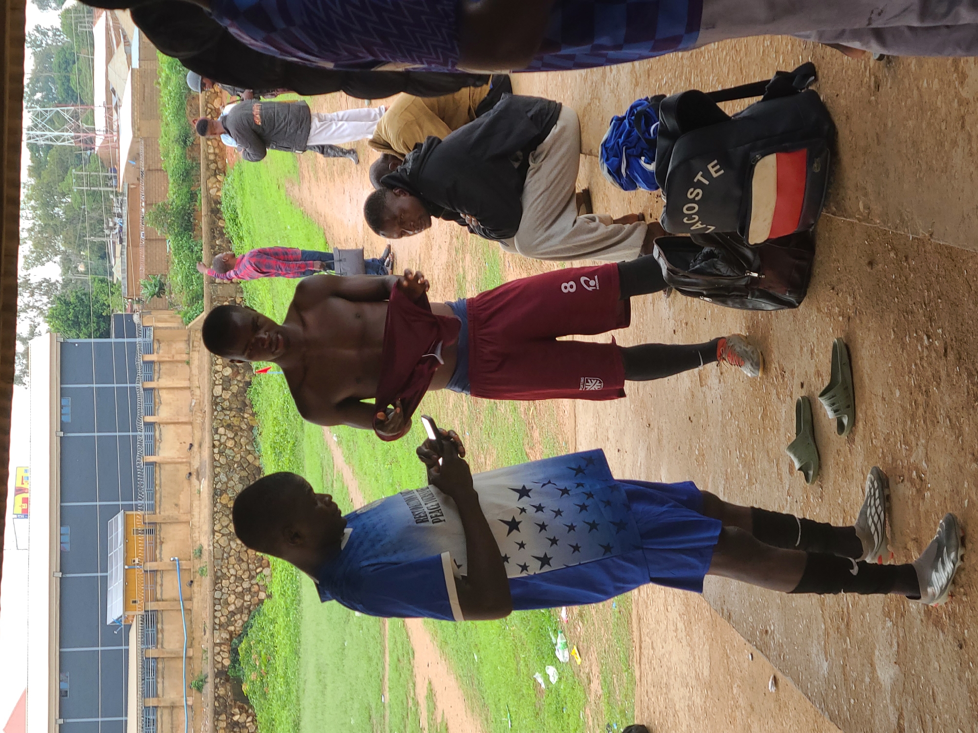 Captured in an outdoor area with a partially visible building in the background, the image portrays a group of young men. One man, shirtless and wearing maroon shorts with the number "8" visible, is holding up a garment. Another man, dressed in a blue soccer uniform with stars, is looking at a mobile phone. Nearby, there is a Lacoste bag and a couple of pairs of shoes. Other individuals are kneeling or standing in the background. The location appears to be in the city of Unknown, Unknown.