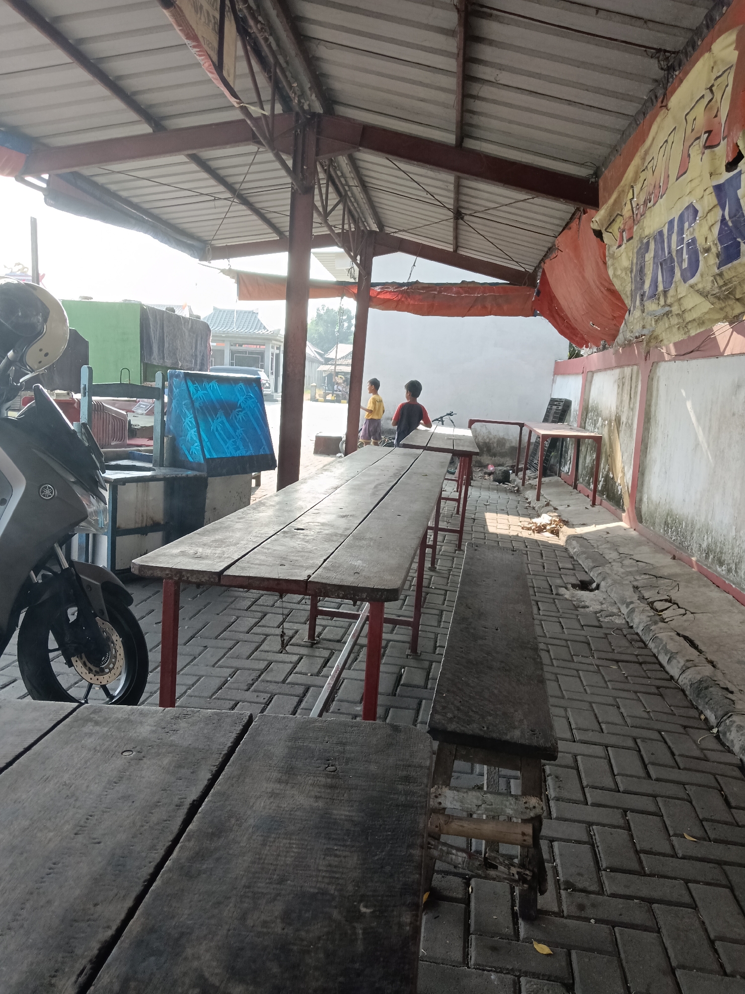 The image depicts an outdoor seating area of what appears to be a roadside eatery or warung in Karawang, Indonesia. Long wooden tables and benches are set up under a corrugated metal roof supported by metal poles. The ground is paved with grey interlocking bricks, and the area shows signs of wear and tear, with some cracks in the paving and peeling paint on the walls. A motorcycle is parked on the left side of the frame, partially obscuring the tables. In the background, two young boys are visible, one in a yellow shirt and the other in a red shirt, standing near a bicycle. The overall atmosphere is casual and utilitarian.