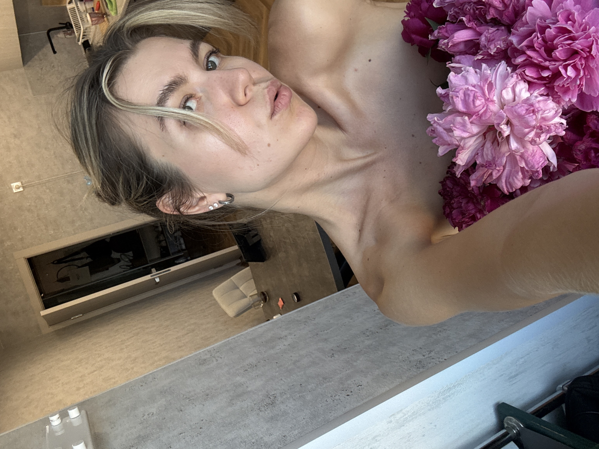 In the foreground of the image, a woman with blonde hair pulled back is holding a bouquet of pink and purple peonies, partially obscuring her chest. She is looking towards the camera with her lips slightly puckered. Her left ear is adorned with a silver hoop earring featuring three small stones. The background shows a domestic interior. To the left, a textured gray wall is visible. Further back, an open doorway reveals a closet with hanging bags and shelves filled with shoes. A dark-colored table with a glass of liquid and some small objects is also present, along with a chair. The lighting suggests it is daytime, with natural light entering from the right side of the frame.