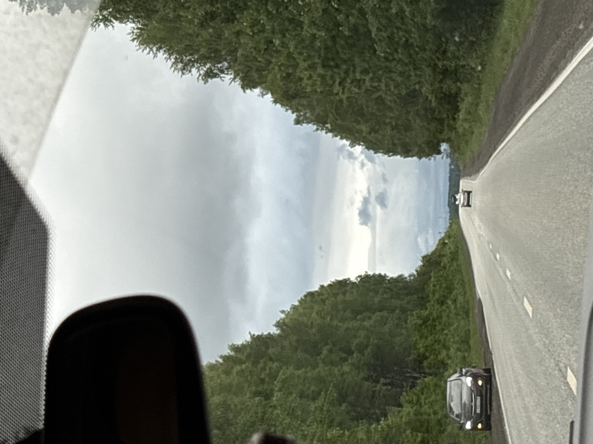 The image shows a view from inside a car, looking out onto a road flanked by trees. The sky is overcast. The road has white lines marking the lanes. There is at least one car ahead on the road and another on the right-hand side of the road. The location is Iiul'skoe, Russia.