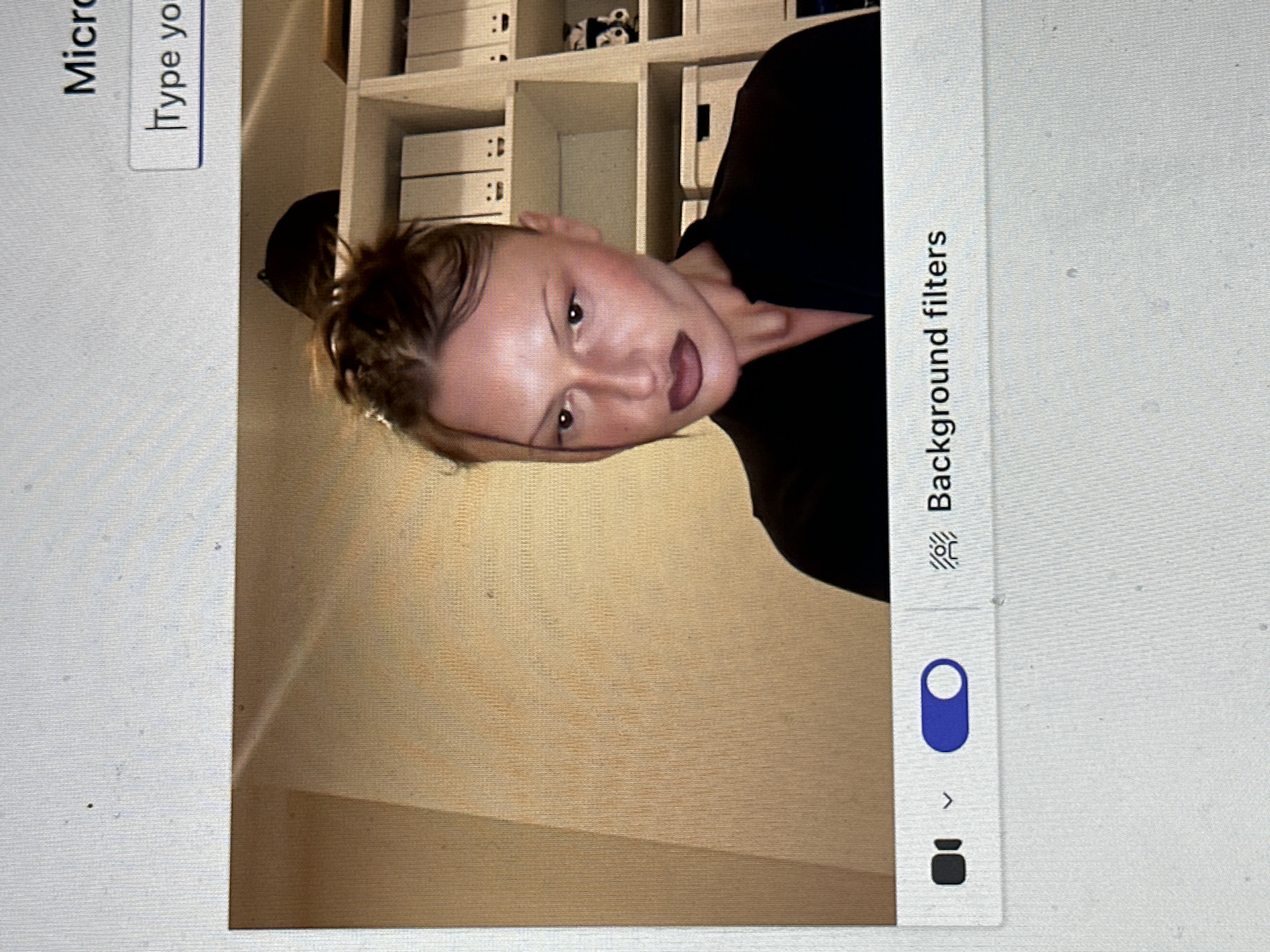 The image displays a computer monitor showing a video feed, likely from a webcam, set within an application interface featuring "Background filters" and "Type yo" text fields. The scene is an indoor setting within Wealden, United Kingdom, though no specific geographical markers are present in the visual content to further pinpoint the exact location.
A female individual is the primary subject, positioned slightly off-center to the viewer's right, facing generally forward with her head slightly angled towards the viewer's left. She has light brown hair styled in an updo, with some strands framing her face. Her facial features include dark eyes, thin eyebrows, high cheekbones, and full lips accentuated with dark lipstick. She is wearing a dark-colored garment, possibly a shirt or blouse, with an open neckline.
The background behind the subject features a light-colored, possibly beige, wall with a distinct angled corner on the viewer's left. To the viewer's right, a white shelving unit is visible, containing multiple white binders or boxes. These binders have black details and a minimalist face-like graphic (two dots and a curved line). A black and white patterned object, resembling a mug, is also on an upper shelf. Partially visible above the subject's head is a dark, rounded object, possibly a cap.
The computer screen itself shows a blue toggle switch in the 'on' position next to a "Background filters" label and icon at the bottom. A camera icon is present on the bottom left. The upper right corner of the screen partially displays the word "Micro" and a text input box labeled "Type yo." The image quality reveals the pixel structure of the display and several small dust particles on the screen surface.