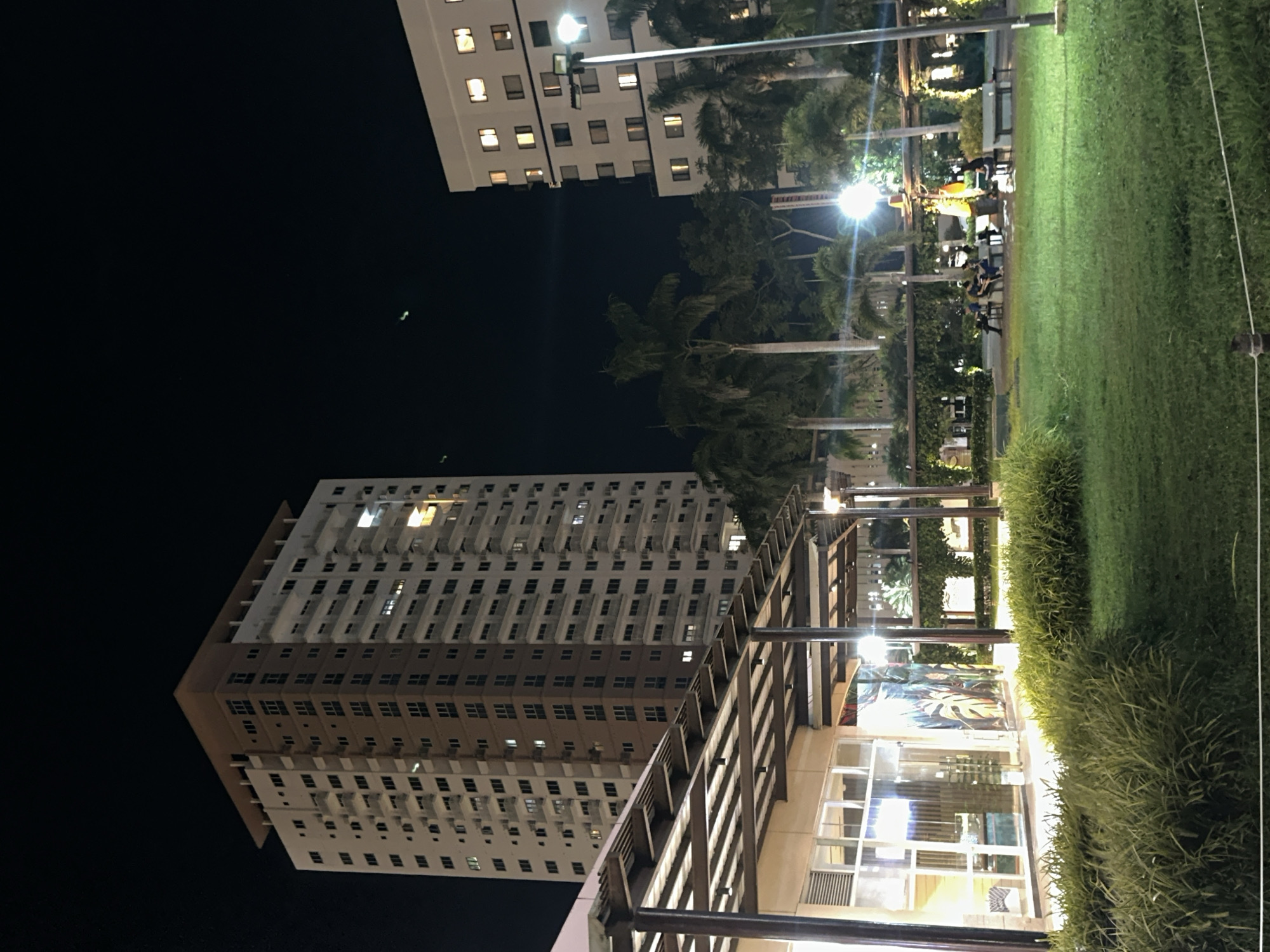 This is a night photograph of an outdoor area adjacent to a tall, multi-story building in Davao City, Philippines. The building on the left is a modern high-rise with numerous windows and balconies. To the right, a shorter, white building with illuminated windows is visible. In the foreground, a manicured lawn with lush green grass is bordered by a pathway and some dense foliage. Several people are seated on benches in a courtyard area, some appearing to be engaged in conversation. There are also palm trees and other vegetation throughout the scene. Artificial lighting from lamps and building lights illuminate the area, creating a contrast with the dark night sky, which features a few faint green lights, possibly aircraft or atmospheric phenomena. A covered walkway or structure with a sloped roof is visible in the mid-ground, supported by pillars.