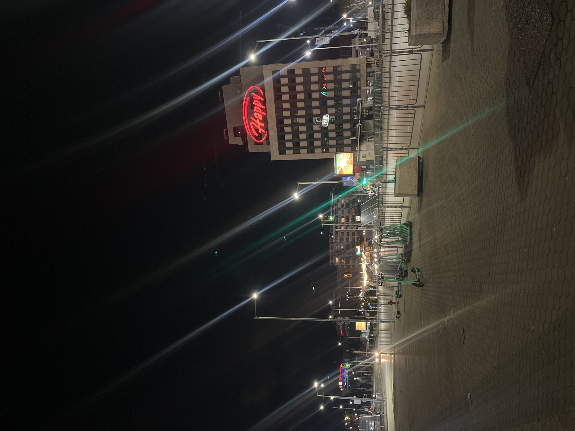 The image depicts a night scene in Sofia, Bulgaria, recognizable by the "Happy" restaurant chain sign illuminated in red on a building. A row of electric scooters is parked on the right side of the frame. Streetlights cast long beams across the scene. The ground is paved with hexagonal bricks. The building with the "Happy" sign has multiple illuminated windows. Other buildings and streetlights are visible in the background. There are metal fences near the sidewalk.