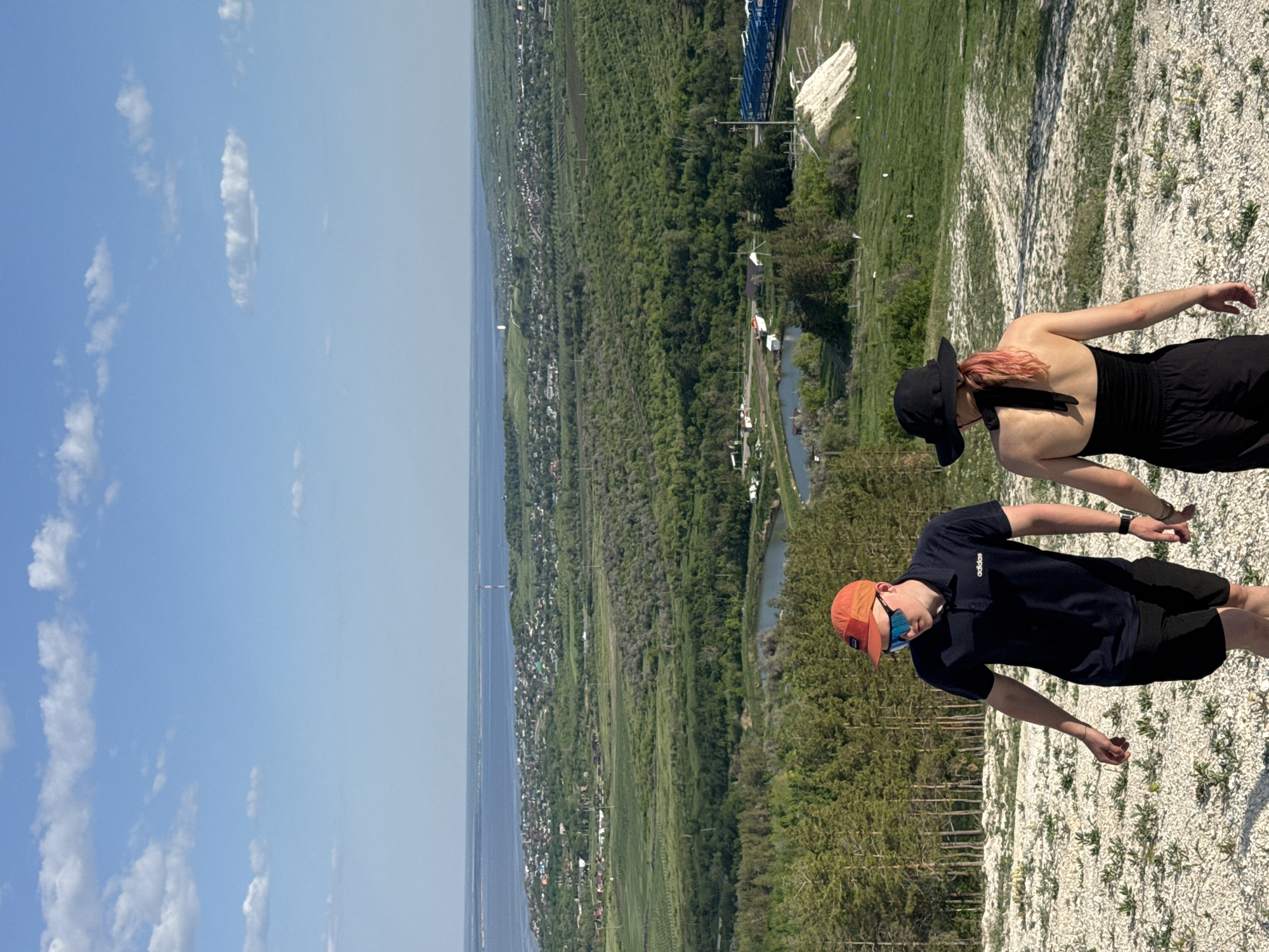 The image depicts a landscape in Khvalynsk, Russia. A body of water stretches along the horizon, bordered by a green expanse, likely representing trees and vegetation. The foreground features two individuals on a rocky, elevated terrain. One person, dressed in black with a black hat, stands behind the other. The second person, wearing a dark blue shirt, black shorts, and an orange cap, is positioned slightly in front. The sky above is blue with scattered clouds. A blue structure is visible in the distance.