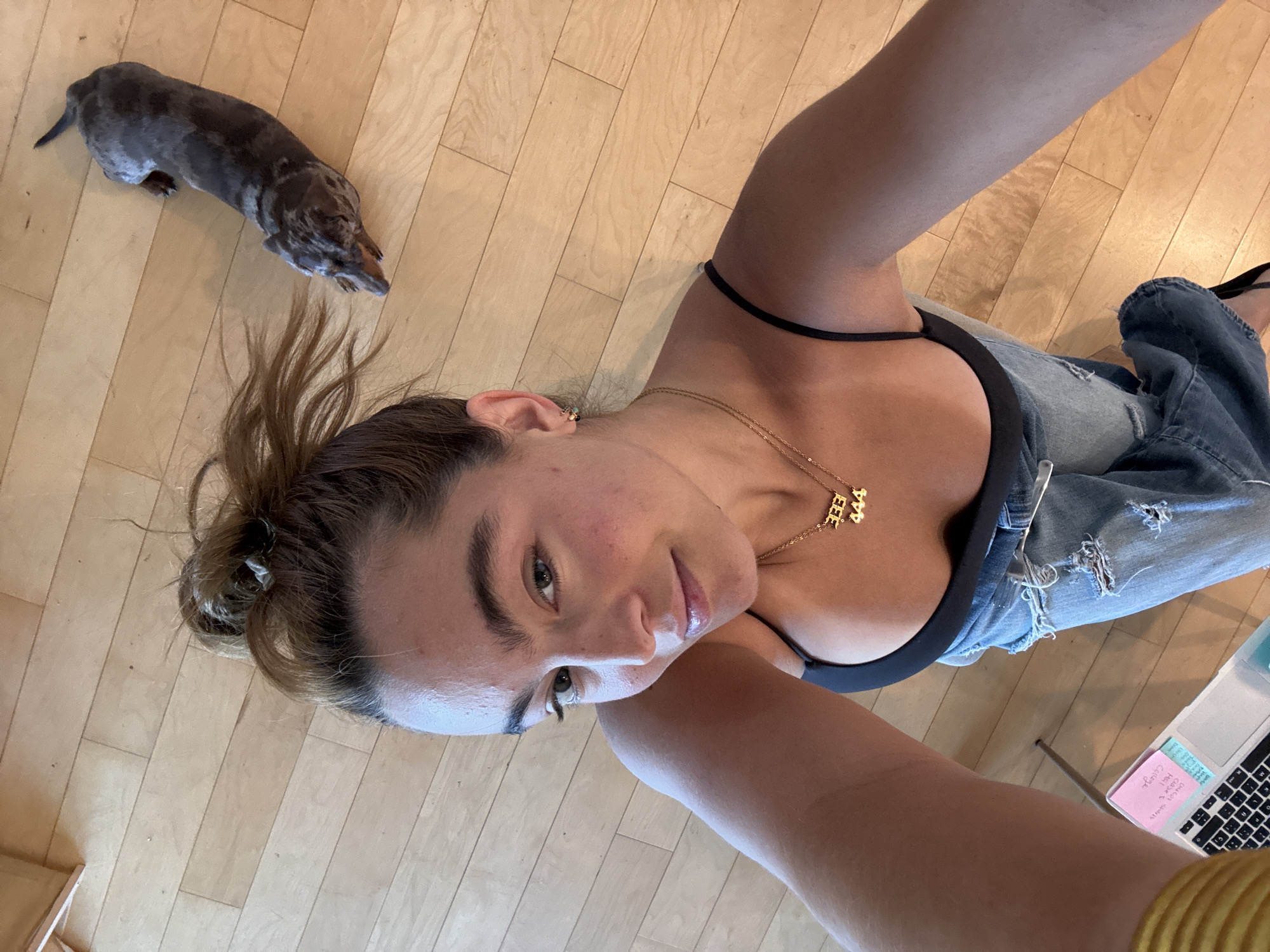 A young woman is depicted in a top-down selfie, lying or leaning on a light-colored hardwood floor in what appears to be an indoor setting in Los Angeles, USA. Her brown hair is partially tied in a messy bun, with some strands framing her face. She wears a black camisole or bralette and light blue distressed denim jeans. A gold-toned necklace with two pendants, one reading "333" and the other "444", is visible around her neck. She has a subtle, soft expression, looking upwards towards the camera.
In the upper left portion of the image, a small, dark-colored dog with a mottled or dapple pattern, likely a dachshund, is resting on the floor, facing away from the camera.
In the lower right corner, a partial view of a silver laptop is visible. Two sticky notes are attached to its screen or keyboard area. The pink sticky note contains handwritten text, partially legible as "chelsea", "Loki", "404 x 3", "macbook", and "magic keyboard". The blue sticky note above it reads "Chicago" and "Hello". The bright, even lighting suggests it is daytime.