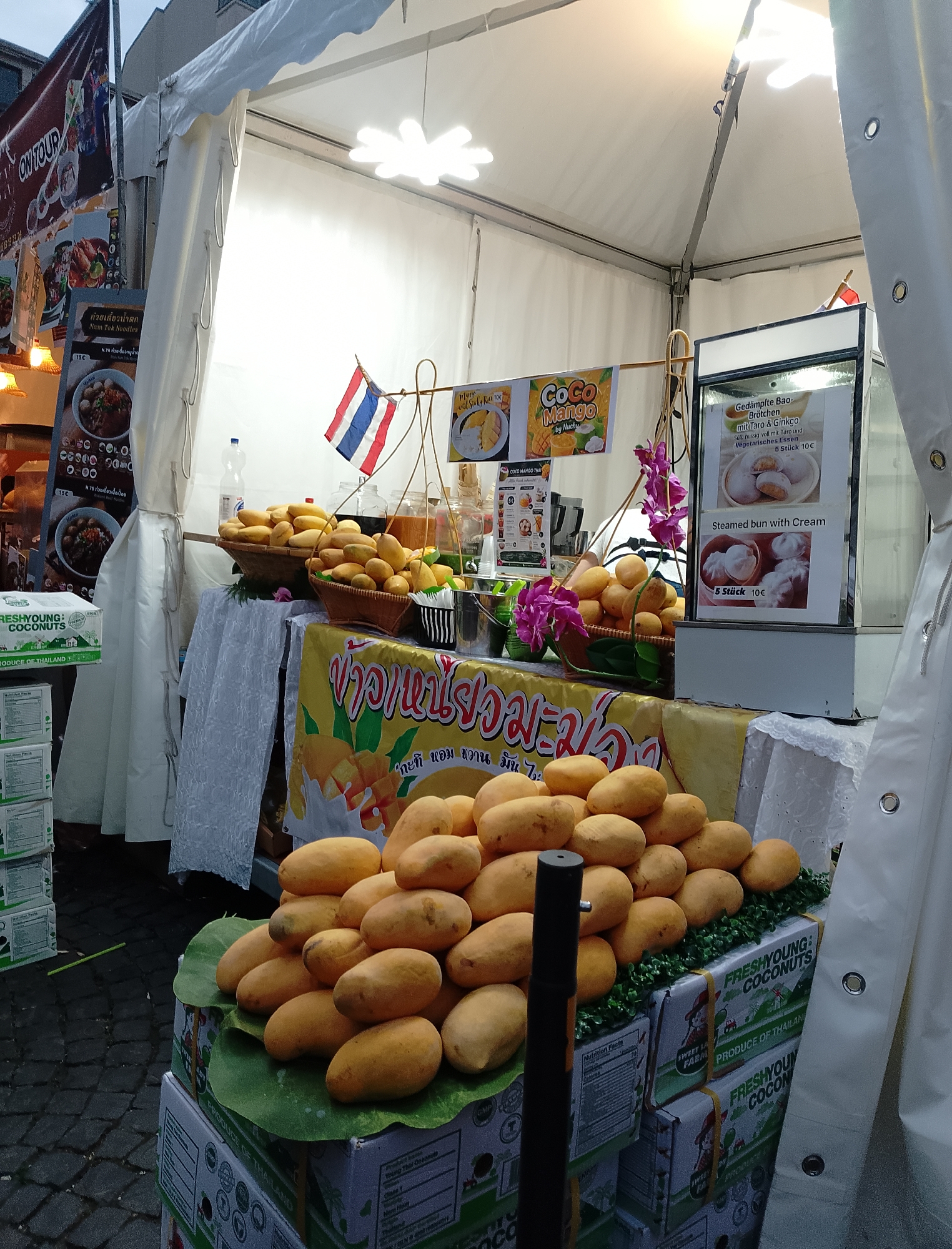 In an outdoor market stall under a white tent, a display of fresh mangoes is prominent, with a large pile of ripe mangoes resting on green leaves atop stacks of "FRESHYOUNG COCONUTS" branded boxes. A Thai flag is visible, and signage in Thai characters suggests Thai cuisine is being offered, with English text indicating "Nam Tuk Noodles" and "Coco Mango." Another sign advertises "Gedämpfte Bao-Brötchen mit Taro & Ginkgo" and "Steamed bun with Cream," priced at 10€ for 5 pieces. The background shows a building and cobblestone pavement.