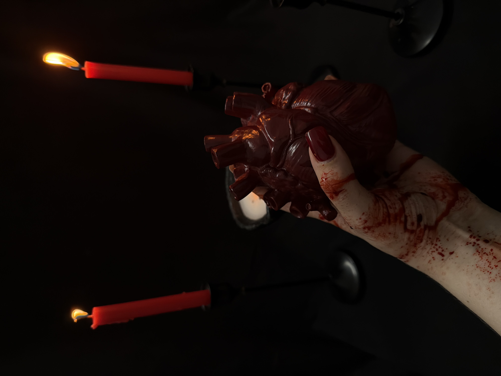 A person's hand, smeared with blood and wearing dark red nail polish, holds a realistic, dark red, anatomical heart model. The scene is set against a black background, with two red candles in black candleholders burning on either side, their flames casting an orange glow. A smaller, white candle is visible in the background. The lighting is dim, with the focus on the hand, heart, and candles. The overall impression is one of a dark, possibly ritualistic, setting.