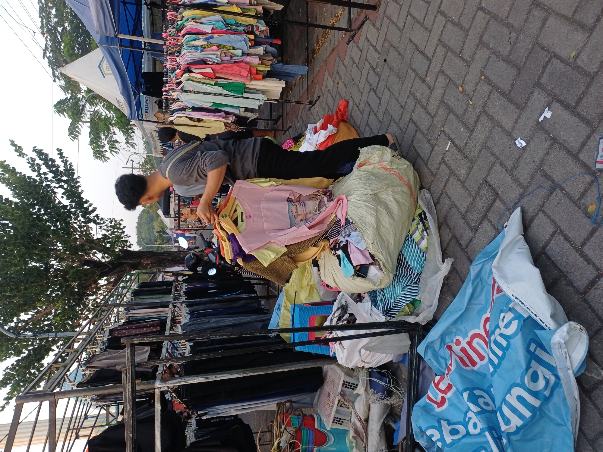 The image depicts an outdoor market scene, likely in Surabaya, Indonesia. A person, viewed from the back, is browsing clothing displayed on racks. The person is wearing a grey long-sleeved shirt, black pants, and dark sandals. They are holding a pink garment with an image on it. In the foreground, a large pile of clothes is visible, with some items in bags and others loose. To the left, a blue tarp with white text is partially visible on the ground. In the background, more clothing racks and market stalls with tents are present. A motorcycle is parked to the right of the clothing racks. The ground is paved with interlocking bricks. The lighting suggests it is daytime, possibly late afternoon given the shadows. The environment appears to be a busy commercial area with a mix of temporary structures and permanent elements like trees and buildings.