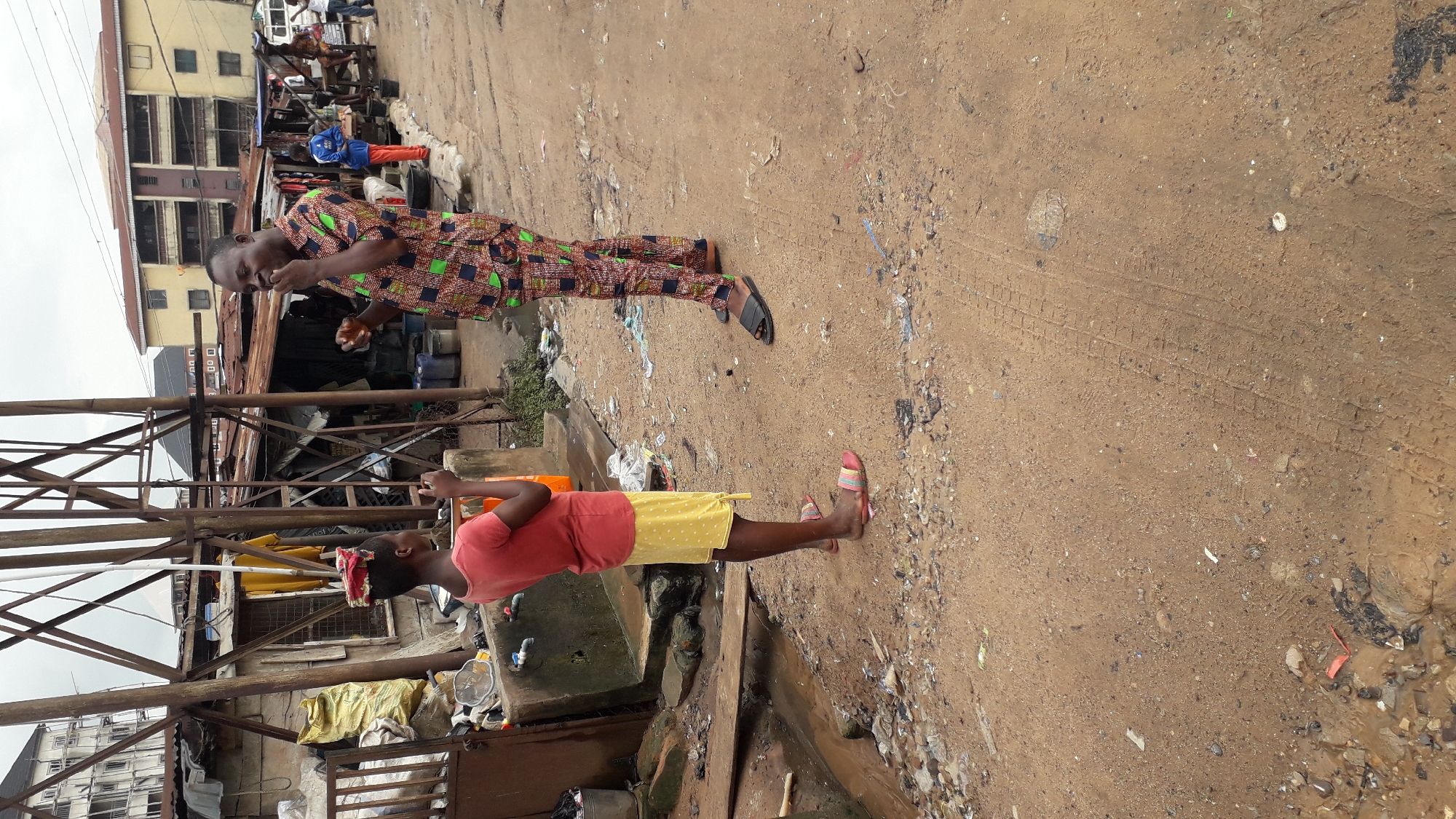 The image depicts a street scene in Onitsha, Nigeria, characterized by its informal urban environment. Two individuals are the central focus. On the left, a young person, possibly a girl, stands with a red and white cloth bundle balanced on their head. They are wearing a coral pink t-shirt and a yellow patterned skirt, with colorful striped flip-flops on their feet. They are looking towards the other individual and holding what appears to be a smartphone.
To the right, an adult male stands facing the younger person. He is wearing a vibrantly patterned short-sleeved shirt with a geometric design in colors like green, black, red, and white, paired with matching trousers. He has dark skin and is smiling, with his mouth slightly open as if speaking or eating something small held between his fingers. He is wearing black slip-on sandals.
The environment is dusty and unpaved, with visible tire tracks in the dirt. In the background, there are makeshift structures made of corrugated metal and wood, suggesting a market or informal settlement area. Some sacks are piled up, and various containers, including blue barrels, are scattered around. Beyond these structures, a multi-story building with yellow and red facades is visible, indicating a more permanent urban presence. The overall impression is one of a busy, typical day in a densely populated, informal urban setting within Onitsha.