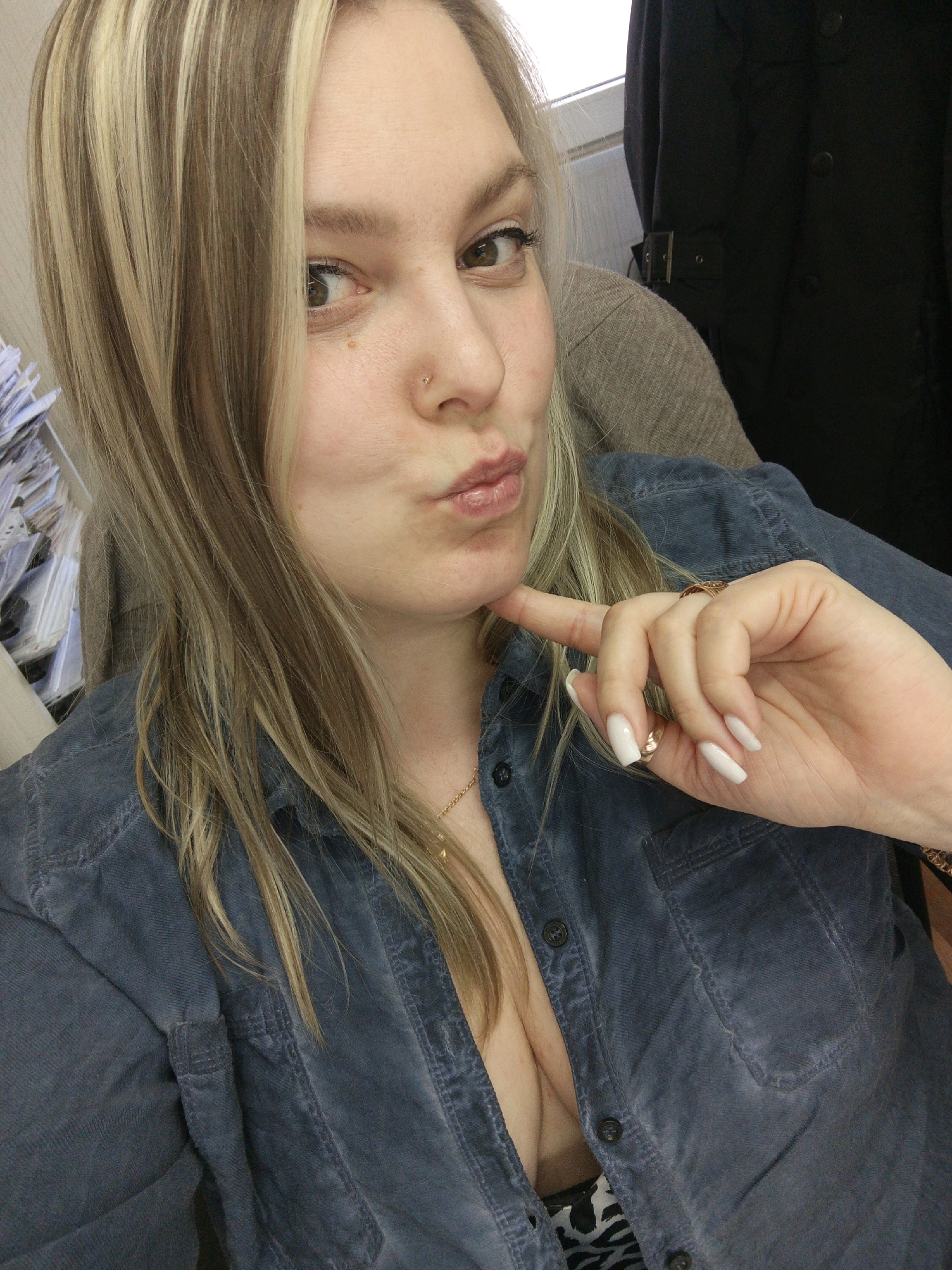 A blonde woman with highlighted hair poses for a selfie indoors. She is wearing a blue button-down shirt and jewelry. Her nails are long and white. She is making a pouting face and touching her chin with her finger. There is a stack of papers to her left and a dark jacket hanging behind her. The location of the photo is unknown, and there is no indication that the photo was taken in Noviy, Russia.