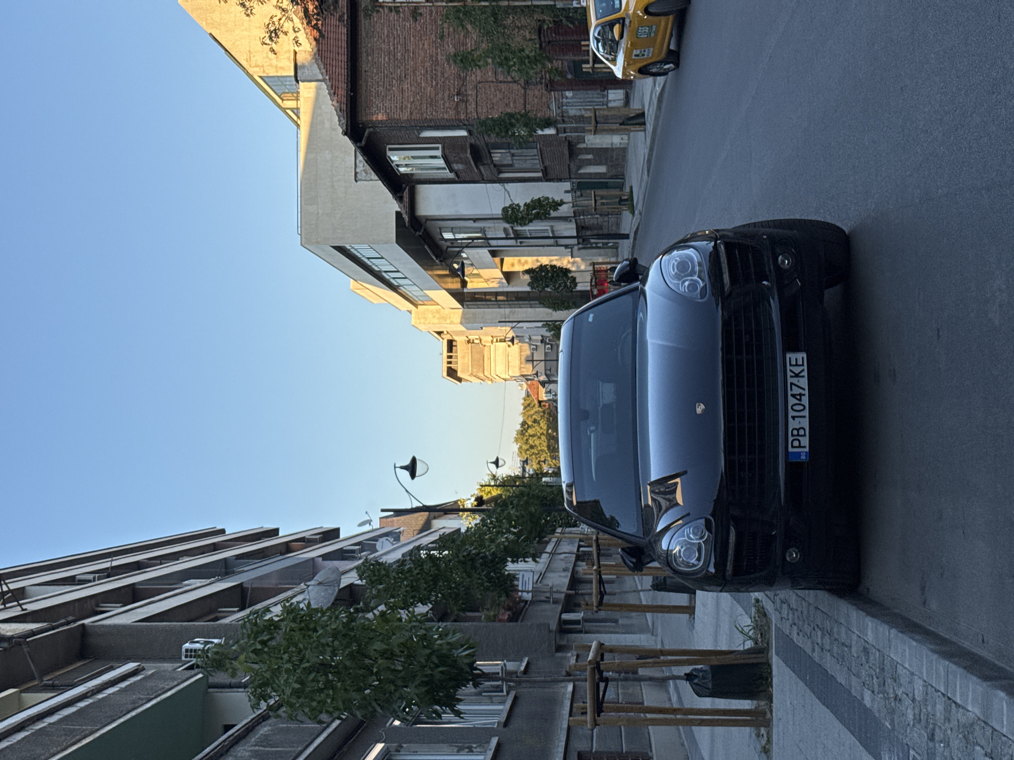A black Porsche Macan is parked on the side of a street in Plovdiv, Bulgaria, with its front end facing the camera. The car is positioned on asphalt, next to a sidewalk lined with young trees supported by wooden stakes. The license plate on the Porsche reads "PB 1047 KE". To the right of the Porsche, a yellow taxi cab is visible further down the street, also parked. The street is lined with buildings on both sides. On the left, a multi-story building with balconies and air conditioning units is prominent. On the right, there are older brick buildings and a more modern concrete structure. The sky is clear and blue, suggesting it is daytime, likely late afternoon or early evening given the warm light casting shadows. There are no people visible in the image. The scene depicts a typical urban streetscape.