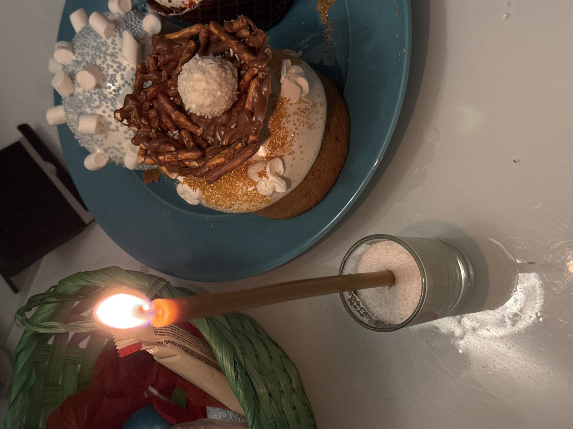A high-angle, close-up shot captures a teal plate with three decorated cupcakes, positioned near a small candle held by a green woven basket. The cupcake decorations include marshmallows, blue sprinkles, chocolate-covered pretzels, gold sprinkles, a white frosted ball, and white frosting flowers. A lit matchstick extends from a glass filled with a white granular substance towards the woven basket. The background is a pale surface. The location is unknown.