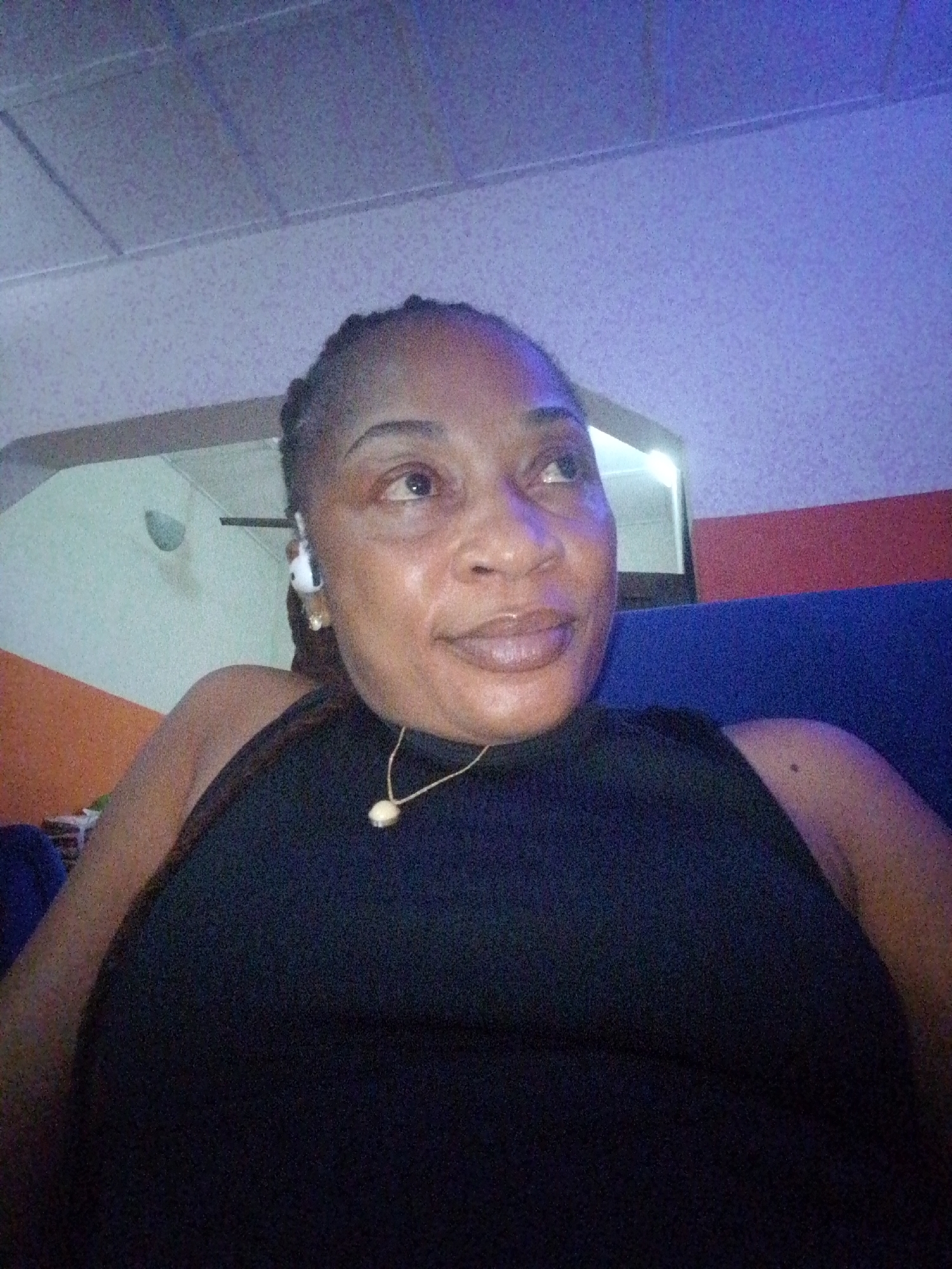 The image is a close-up, waist-up selfie of a Black woman with braided hair, wearing a sleeveless black top and a delicate necklace with a pendant. She is wearing white wireless earbuds. The woman is looking upwards and slightly to her right, with a faint smile on her lips. The lighting is dim and casts a blue and purple hue on her skin and the background, which appears to be an indoor setting with a textured ceiling. The background also shows a wall with orange and light green sections and a dark blue surface, possibly a couch. There are no clear indications of the location within a city, or any specific environmental context that would suggest a city.