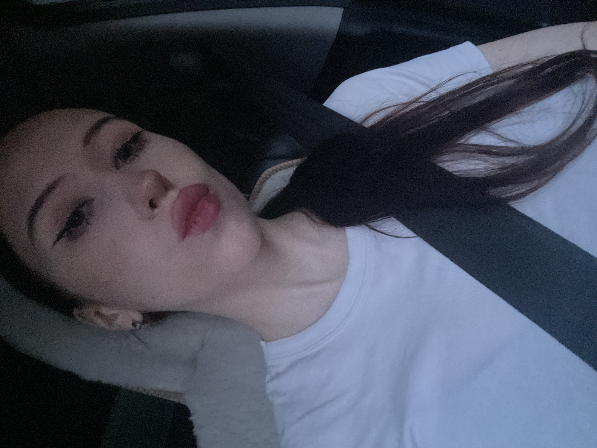 A young woman with dark hair, wearing a white t-shirt, poses for a selfie inside a vehicle. She has on eyeliner and lip gloss and is making a kissing face. A seatbelt is visible across her chest. The background is dark, with a white fluffy headrest visible to her left. The image appears to be taken from inside the car, looking towards the driver or passenger seat. There is no indication of the city of Votkinsk, Russia, or any specific location.