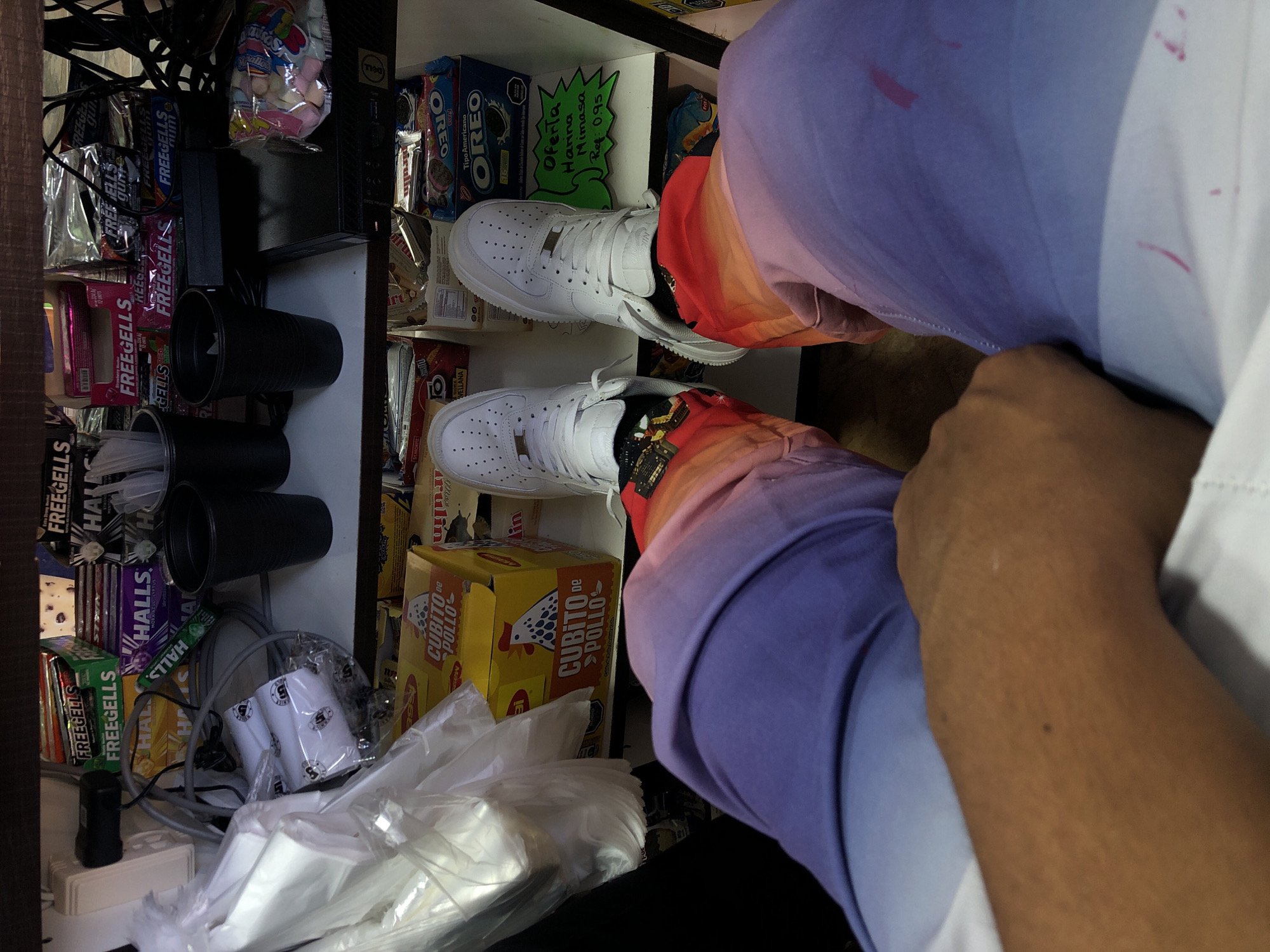 The image shows a pair of white Nike Air Force 1 sneakers worn by a person with colorful socks and ombre-colored pants, apparently sitting in front of a shelf full of snacks and supplies. The shelf contains items such as Oreo cookies, Halls cough drops, Freegells gum, Cubito de Pollo bouillon cubes, and other packaged goods. There are also black cups, plastic-wrapped items, and various electronic cables visible on the shelf. The image is taken from an overhead perspective. It is not possible to identify the location in Juan Griego, Venezuela from this image alone.