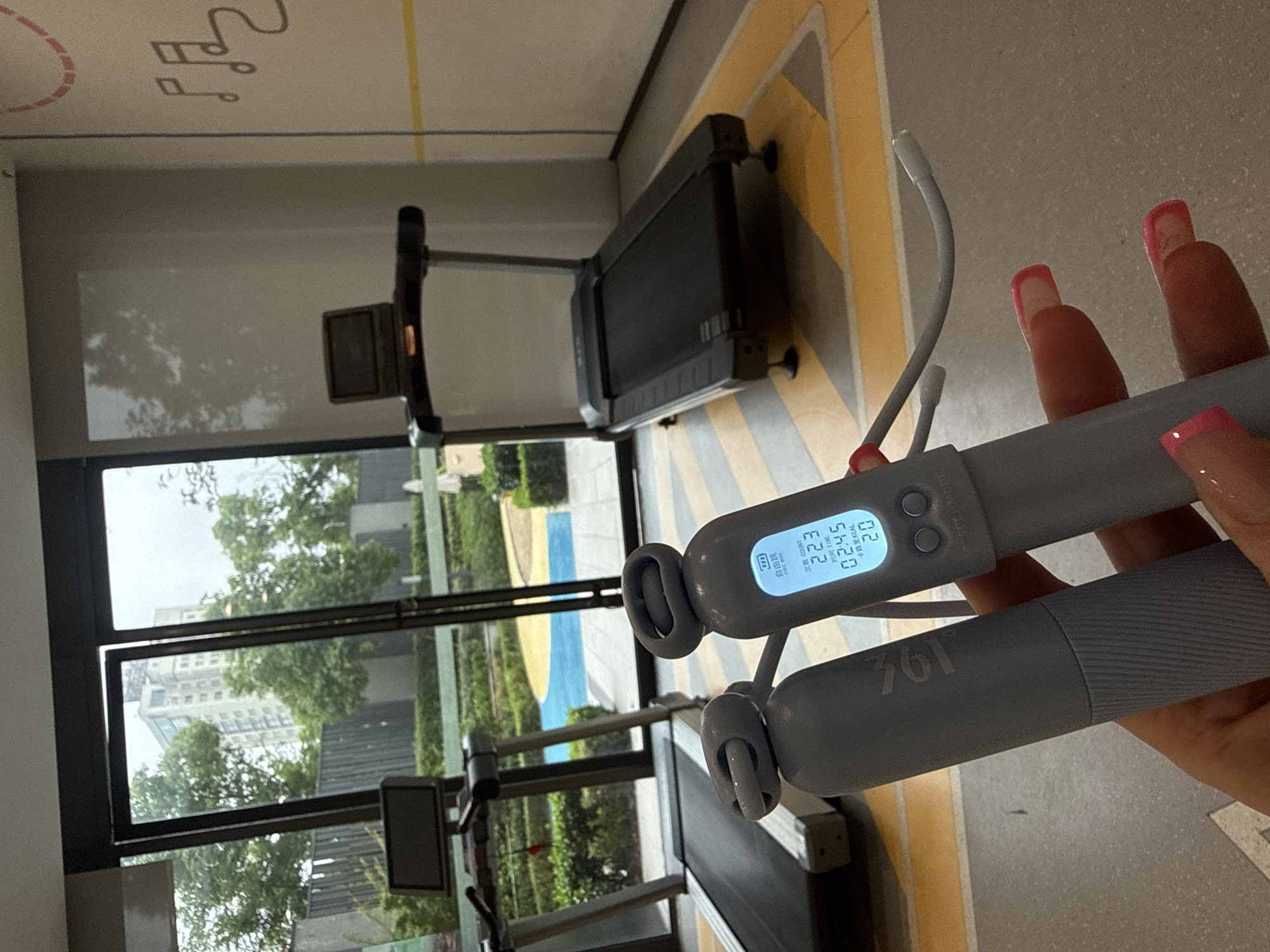 The image captures an indoor fitness setting, likely a gym or exercise area in Yiwu, China, seen through the perspective of a person holding a smart jump rope. The main subjects are a person's hand with pink-tipped manicured nails, gripping a grey digital jump rope, and the interior of the fitness space with large windows revealing an outdoor scene.
The smart jump rope's display is illuminated, showing workout statistics. Visible text on the screen includes "自由跳" (Free Jump), "FREE WIXE" (likely a stylized "FREE JUMP"), "次数 COUNT" with a value of "223", "时间 TIME" showing "02:45", and "卡路里 KCAL" with "20". This indicates an ongoing or recently completed jump rope session. One handle of the jump rope is marked with "361", suggesting a brand name. The jump rope appears to have a traditional rope attached, but also a ropeless option with small weighted balls.
The background shows two treadmills positioned along a wall with a floor marked by yellow and grey geometric patterns. Large windows span the wall, offering a view of lush green trees and modern buildings outside under what appears to be a bright, clear daytime sky. The setting suggests a well-lit and comfortable environment for exercise.