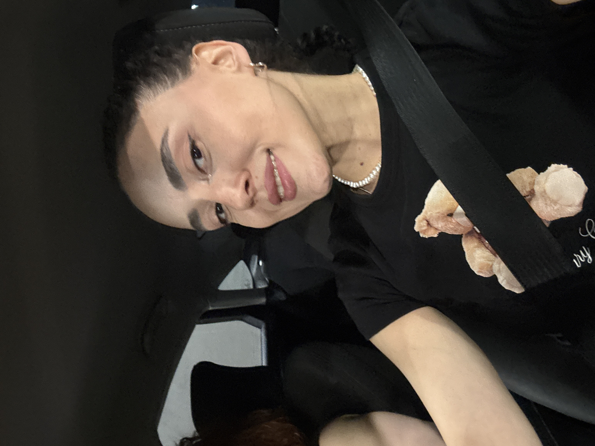 A young woman wearing a black t-shirt with a teddy bear graphic and a diamond necklace is in the passenger seat of a car. She has dark hair pulled back, dark eyebrows, and light skin. She is smiling slightly and looking at the camera. A seatbelt is across her chest. The car interior is dark. The location is unknown and not discernible from the image.