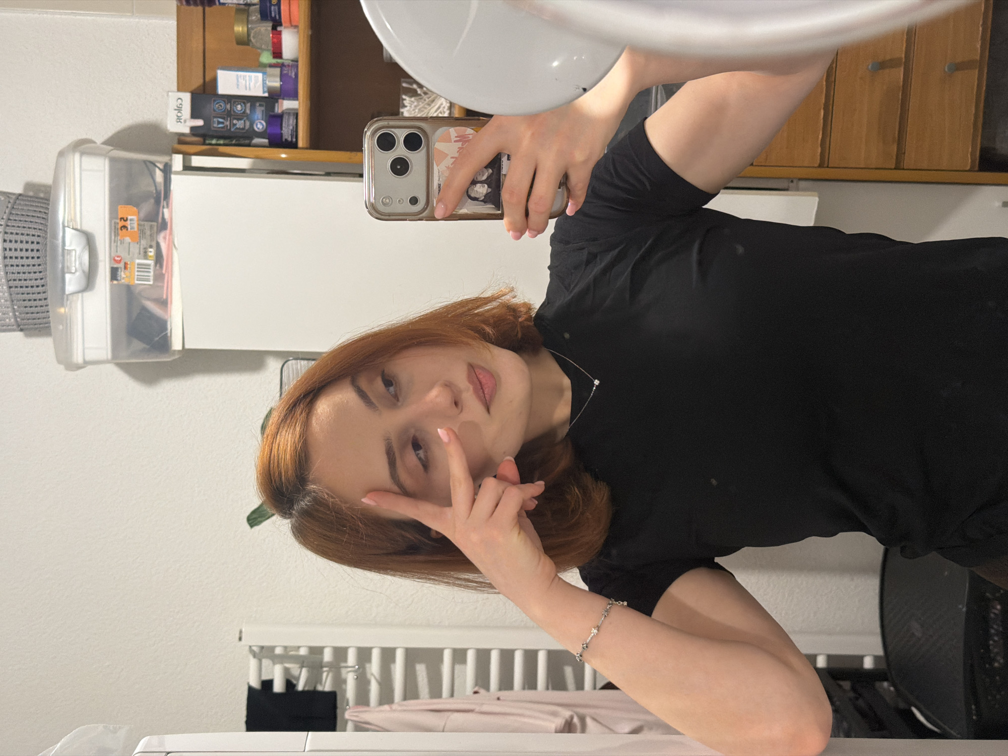 The image shows a young woman with auburn hair taking a selfie in a bathroom. She is wearing a black t-shirt and a silver bracelet. She is holding a smartphone in her right hand and has her left hand raised to her forehead, making a peace sign. Her hair is shoulder-length and styled with a slight wave. The background includes a white wall, a white radiator with pink clothing hanging on it, a shelf with various items including toiletries and a transparent storage box, and a wooden dresser. The woman appears to be in a private residence, and there is no indication of any specific location within the city of Strasbourg, France.