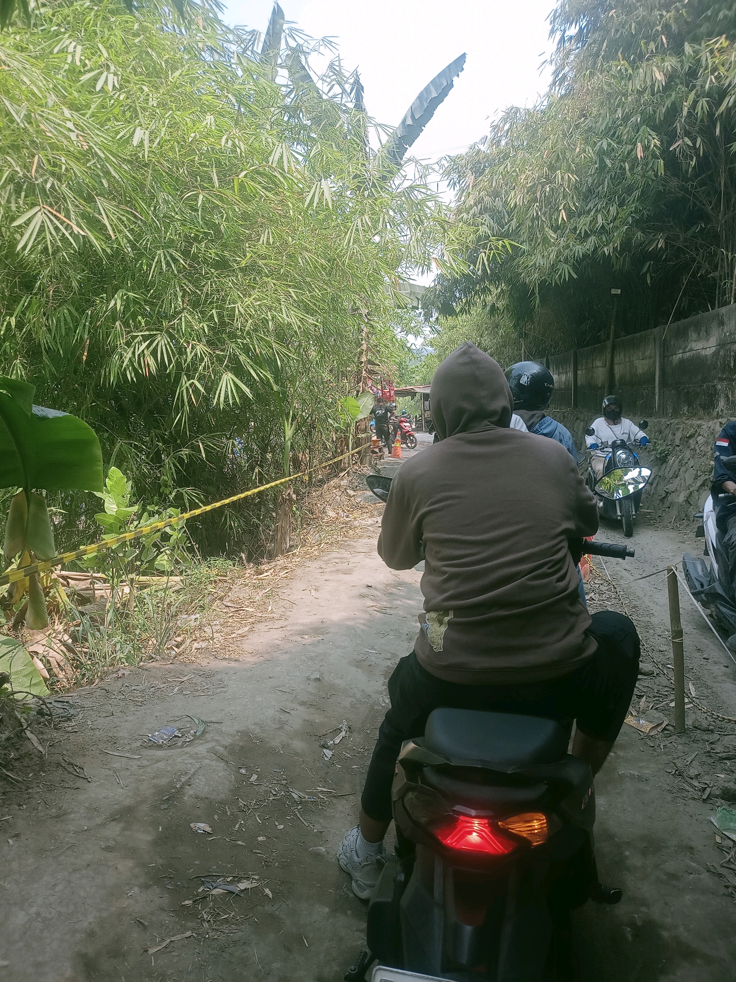 The image captures a sunny day scene in Klapanunggal, Indonesia, likely along a rural or semi-rural road. A person wearing a brown hooded sweatshirt is seated on the back of a black scooter, facing away from the camera. The scooter's brake light is illuminated. Beside this scooter, another scooter is visible, and further down the road, at least two other scooters with riders are present, one with a rider wearing a white long-sleeved shirt and a dark helmet, and another with a rider in a blue shirt and dark helmet. The road appears to be made of dirt or gravel and is lined with dense bamboo foliage on the left and a concrete wall on the right, suggesting it's a pathway through a vegetated area. Yellow caution tape is strung across the path on the left side, partially obscured by a large green leaf. The ground is littered with dry leaves and debris. The overall impression is of a simple, everyday scene of people traveling by scooter through a natural environment in the specified location.