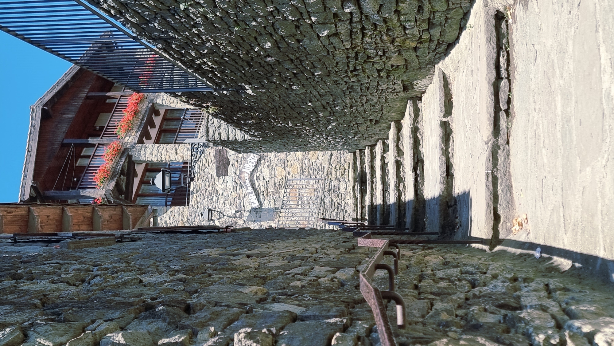 The image depicts a narrow, cobblestone alleyway in the town of Rupit i Pruit, Spain. Stone walls flank both sides of the alley, with a steep staircase made of large, flat stones leading upwards. To the left, a weathered metal handrail runs along the staircase. On the right, a portion of a building is visible, constructed with stone and wood. This building features balconies adorned with vibrant red flowers and a dark wooden facade. A sign is mounted on the stone wall, partially obscured, with text visible that appears to read "BAR RESTAURANT ALBER..." indicating it is a dining establishment. The sky above is a clear, bright blue, suggesting a sunny day. The overall scene conveys a sense of rustic charm characteristic of historic European villages.