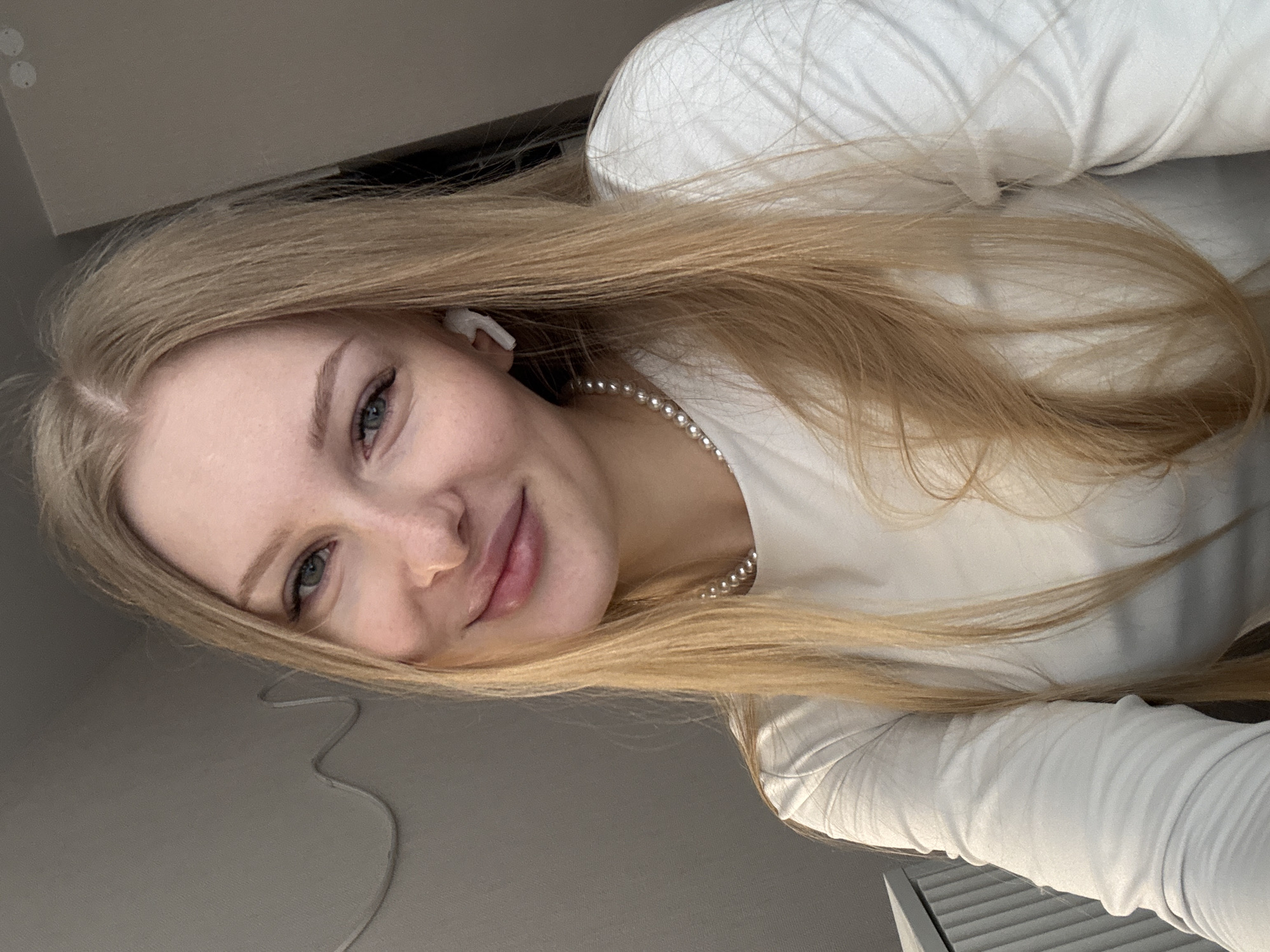 A young woman with long, straight blonde hair and light skin is featured in a close-up, upward-angled shot. She is smiling softly, looking directly at the camera with her green-grey eyes. She wears a simple white long-sleeved top and a pearl necklace. A white earbud is visible in her right ear. Her expression is relaxed and pleasant. The image is rotated 90 degrees clockwise, suggesting she might be lying down or leaning back. The background is softly blurred, showing an indoor setting with muted, light grey tones and what appears to be a ceiling or wall, along with some darker, indistinct elements to the right. There is no visible text. The lighting is soft and even, suggesting an indoor daytime setting without harsh direct light.