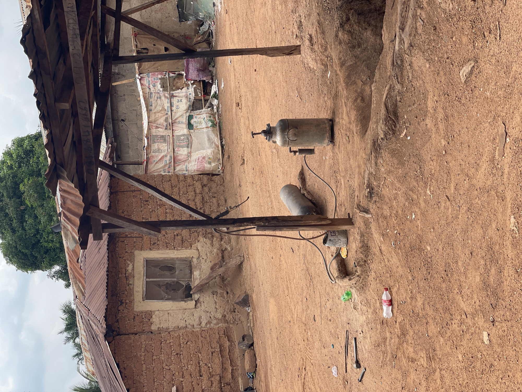 The image captures an outdoor scene, likely a yard or open workspace, in what appears to be a rural or semi-urban setting. The ground is a reddish-brown, uneven dirt surface, typical of many African landscapes, with small rocks and some debris scattered about.
To the left, a rustic building features walls constructed from mud bricks or rammed earth, showing signs of wear and repair. Its roof is made of corrugated metal sheets, some appearing weathered and slightly damaged, supported by rough-hewn wooden beams. A single window in this building has broken or missing panes. Green foliage from trees, including what looks like a mango tree, is visible above and behind this structure, against a partially cloudy sky.
In the background, a more open shelter or partially constructed building is visible. Its walls are composed of stacked white sacks, clearly labeled "DANGOTE CEMENT" with "3X" and "RELIABLE PRODUCT" visible on multiple bags, indicating they are likely used as temporary walling or for storage. This structure also has a wooden support frame.
The foreground and central area feature several objects on the dirt ground. A prominent, metallic, cylindrical tank, possibly a large propane or LPG tank, stands upright with a valve on top. A hose extends from it to a smaller, grey cylinder lying on its side. Nearby, various tools and scraps of metal are scattered, including pieces of rebar and what resembles a hammerhead. A red-capped plastic bottle, possibly a soft drink bottle, and a green plastic container are also on the ground. To the far right, the ground shows a slight depression or eroded area.
No people are present in the image, but the arrangement of tools, gas tanks, and cement bags suggests an area used for work, possibly construction, repair, or a small-scale workshop. The lighting indicates it is daytime, under a bright but somewhat overcast sky. The overall impression is one of a functional, utilitarian space in Akwanga, Nigeria.