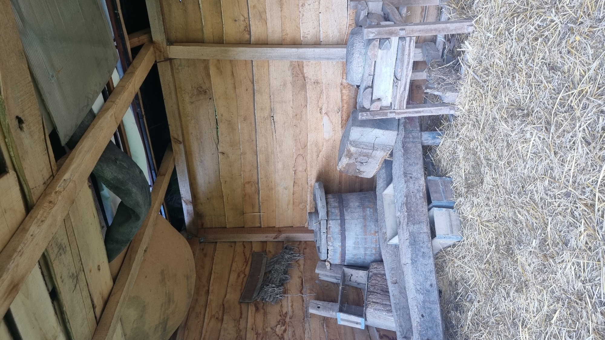 The image displays the interior of a rustic wooden structure, likely a barn or shed, located in Melnyky, Ukraine. The walls are constructed from horizontal, unpainted wooden planks exhibiting wear and varying hues. The ceiling features exposed wooden beams and planks, with gaps revealing a darker area beyond and draped fabric or netting. A corrugated metal sheet is also visible in the upper right ceiling section.
The dirt floor is covered with a thick layer of dry straw or hay. No people are present.
Various aged wooden objects and tools are distributed throughout the scene. On the upper left wall, a metal or leather strap secures a bundle of coarse ropes or netting. On the floor, a large wooden barrel with metal bands is centrally located, topped with a circular wooden object, possibly a wheel component or a lid. To the left of the barrel, a large wooden log or stump supports a light blue, open wooden frame. Several large, roughly hewn wooden beams or timbers are laid horizontally across the straw. To the right, a rudimentary wooden frame, resembling a chair, holds several round, flat stones, suggestive of grinding stones. Additional rough wooden blocks and small wooden boxes are partially visible under the horizontal timbers and amidst the straw. The environment appears to be an aged, utilitarian storage or workspace.
