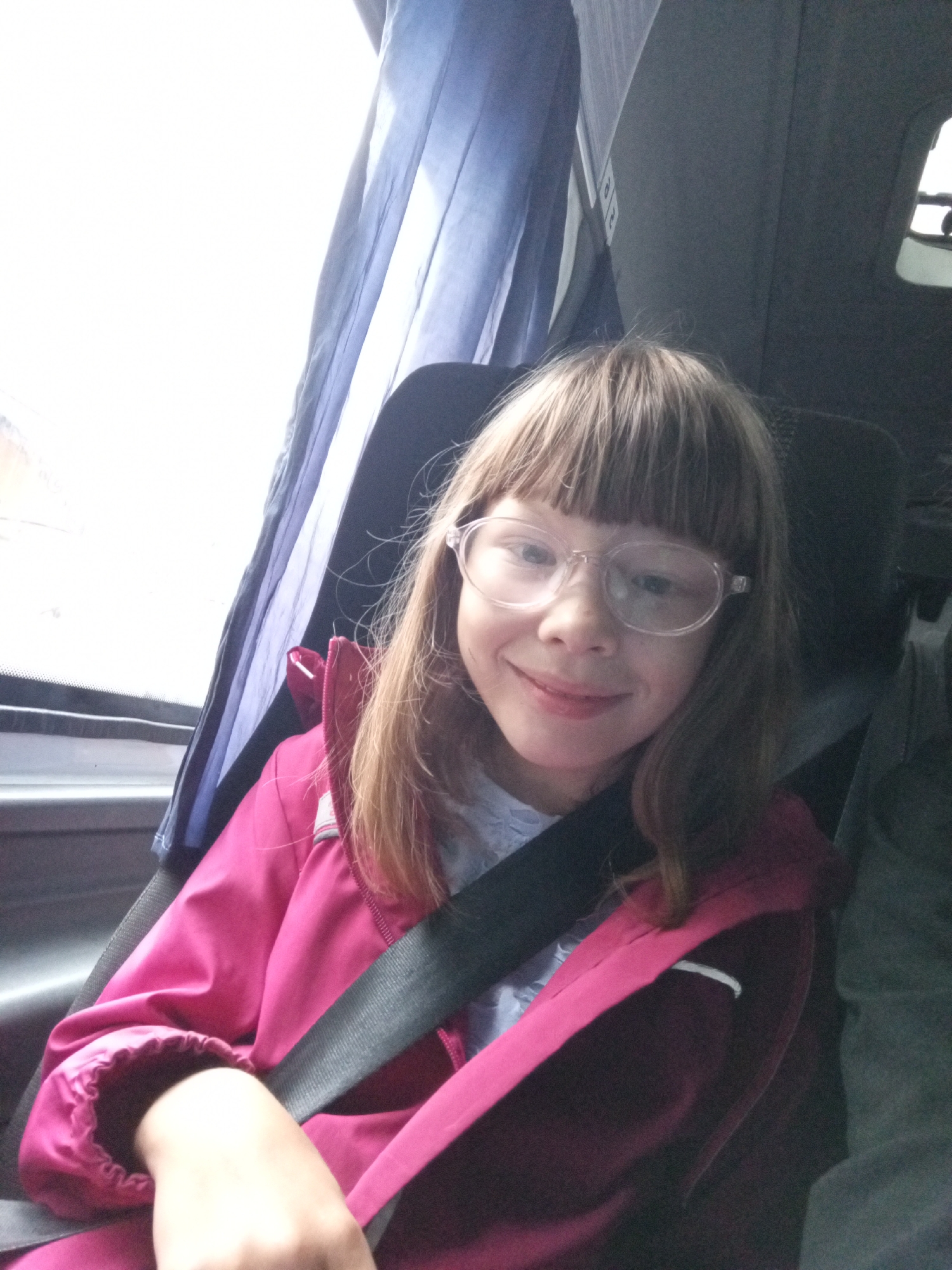 In Verkhneyarkeyevo, Russia, a young girl with blonde hair and bangs, wearing glasses and a pink jacket, sits in a bus seat. She is wearing a seatbelt and looking at the camera with a slight smile. The bus interior is visible, with blue curtains to the left and a dark gray seat behind her. The lighting suggests it is daytime.