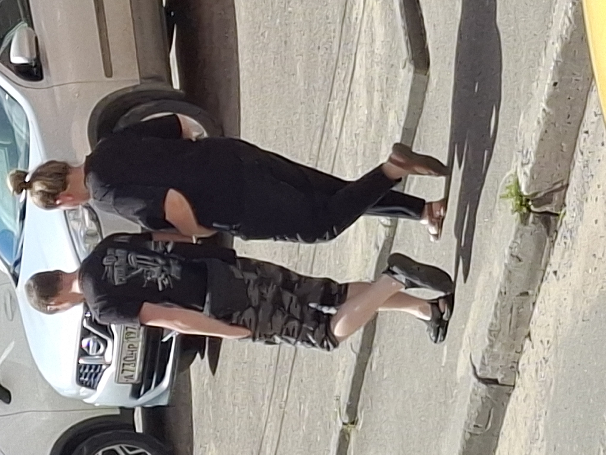 Two individuals, a male and a female, are walking on a paved surface next to a light-colored car. The male, on the left, is wearing a black t-shirt with a graphic design and camouflage shorts. The female, on the right, is wearing a black t-shirt and black pants. She has her hair pulled up in a bun. They are walking in the same direction, away from the viewer, casting shadows on the ground. The environment appears to be an urban setting, with a concrete curb in the foreground and the car parked behind the individuals. The license plate of the car is partially visible, showing characters like "A770HP 197".