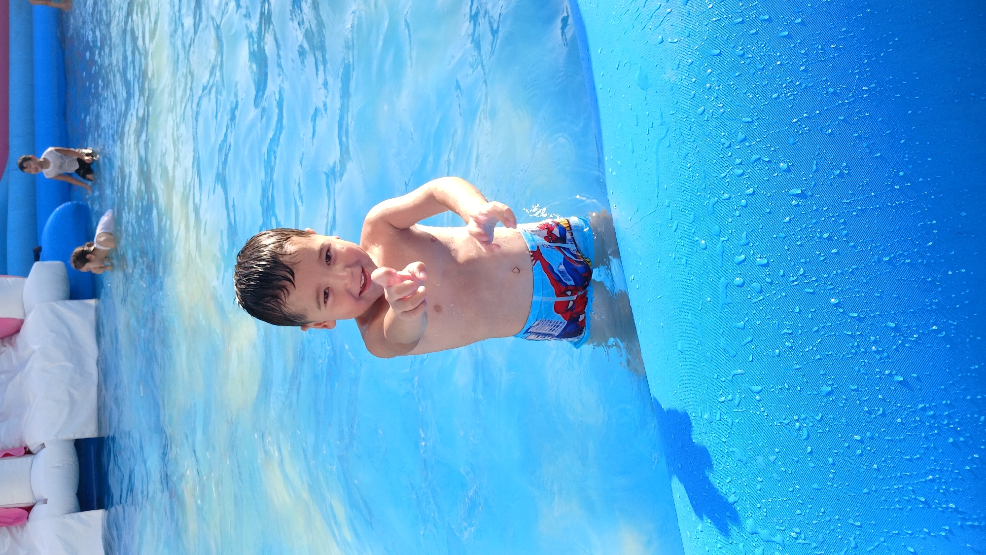 A young boy with dark, wet hair and a wide smile stands in a shallow, bright blue swimming pool. He is wearing blue swimming trunks with a Spiderman print. His body is partially submerged in the water, and he is pointing his right index finger directly at the camera with his left hand slightly extended. Water droplets are visible on his skin. In the background, other children are visible in the pool, along with a large inflatable structure with white and pink sections. The water is clear and reflects the bright sky. The bottom of the pool appears to be a solid blue surface, possibly an inflatable liner. The overall scene depicts a day of recreation at an outdoor water park or pool.