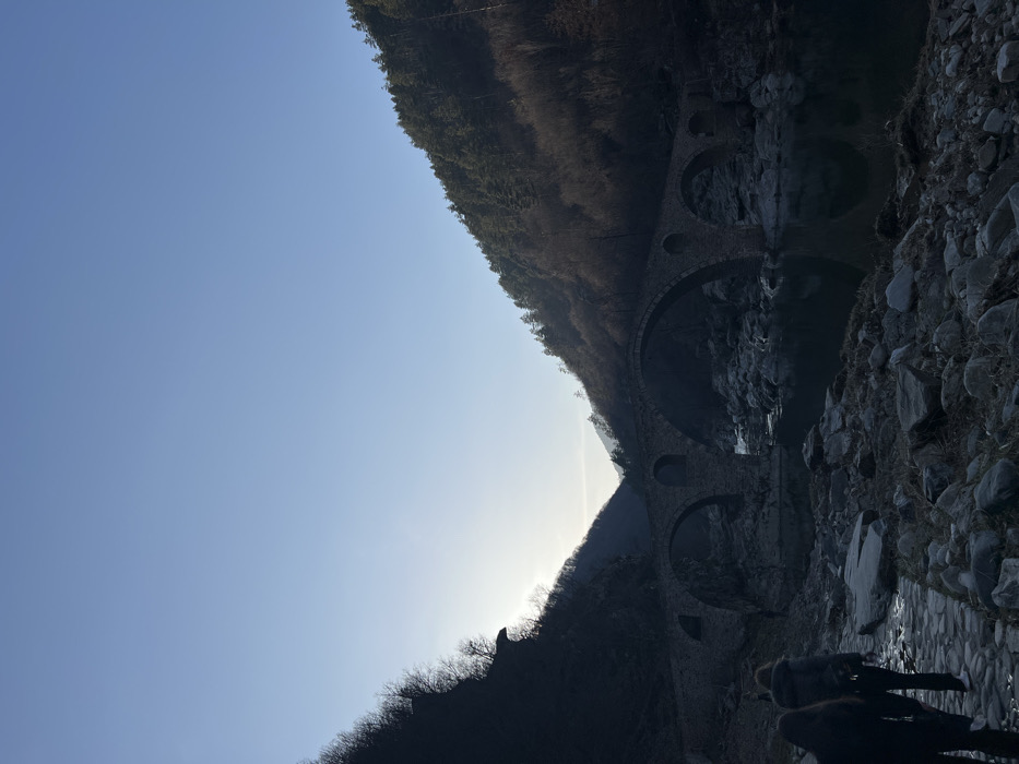 The image depicts a stone arch bridge, identified as the Devil's Bridge (Dyavolski most), situated over the Arda River in a forested mountain valley near Diadovtsi, Bulgaria. The sky above is clear and light blue, with the sun positioned to the upper left, causing backlighting across much of the scene. Two individuals, dressed in dark clothing, are walking away from the camera on a cobblestone path in the lower-left foreground, moving towards the bridge. The river below the bridge contains dark, reflective water flowing over a rocky bed with numerous exposed stones. Steep, tree-covered hillsides frame the valley, with the left slope appearing darker due to shadow and the right slope showing more direct sunlight on its evergreen and deciduous trees. The bridge itself is constructed of dark grey stone, featuring multiple prominent arches and characteristic circular relief holes.