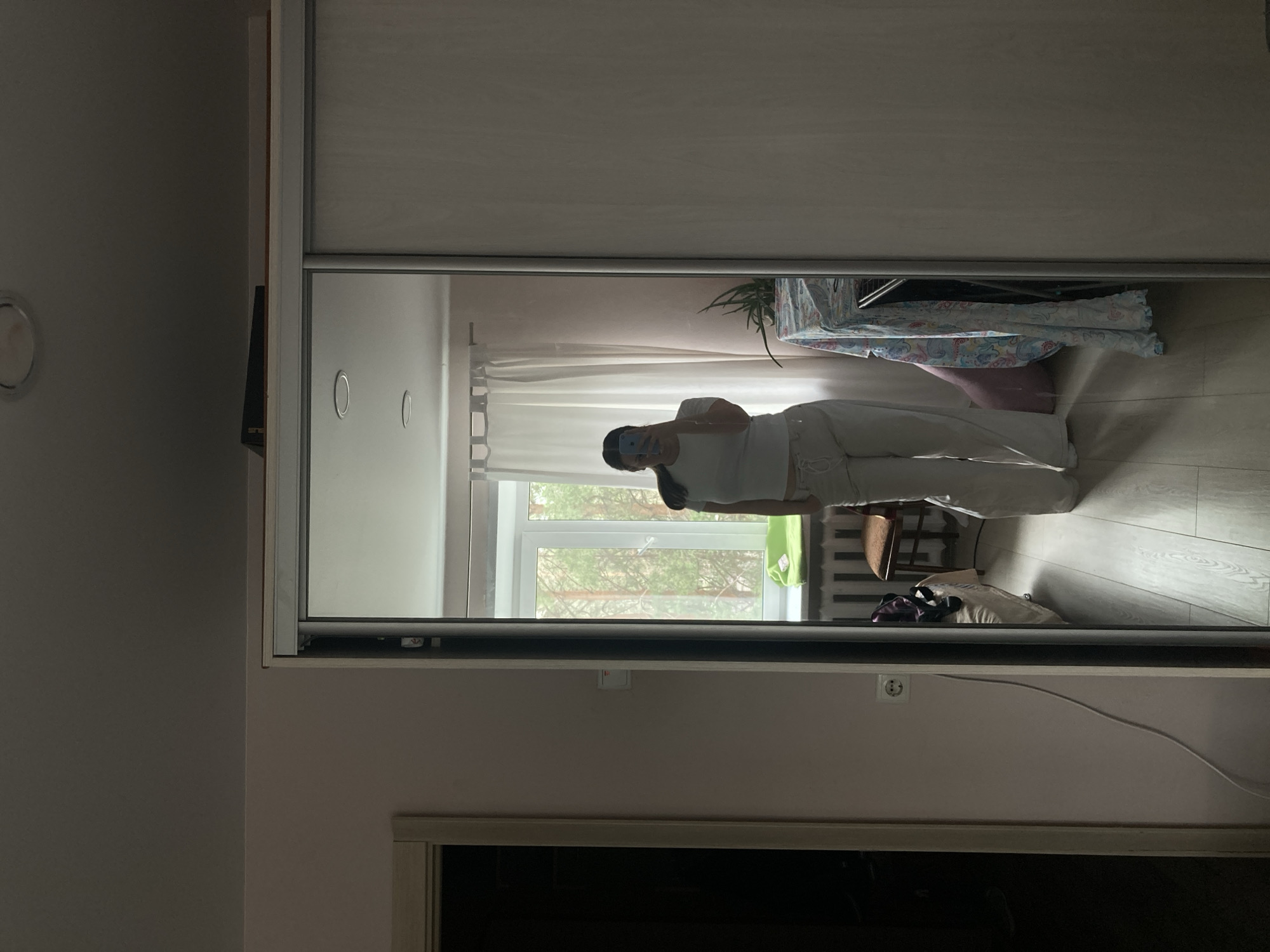 A mirrored closet door reflects a room in Chaikovsky, Russia. A person holds a phone, capturing the scene in the mirror's reflection. The room contains a window with visible trees outside, a table covered with a floral cloth, a chair, a radiator, and clothing on the floor. The walls are painted in a light shade of pink, and there is an electrical outlet visible near the bottom of the mirror's frame.