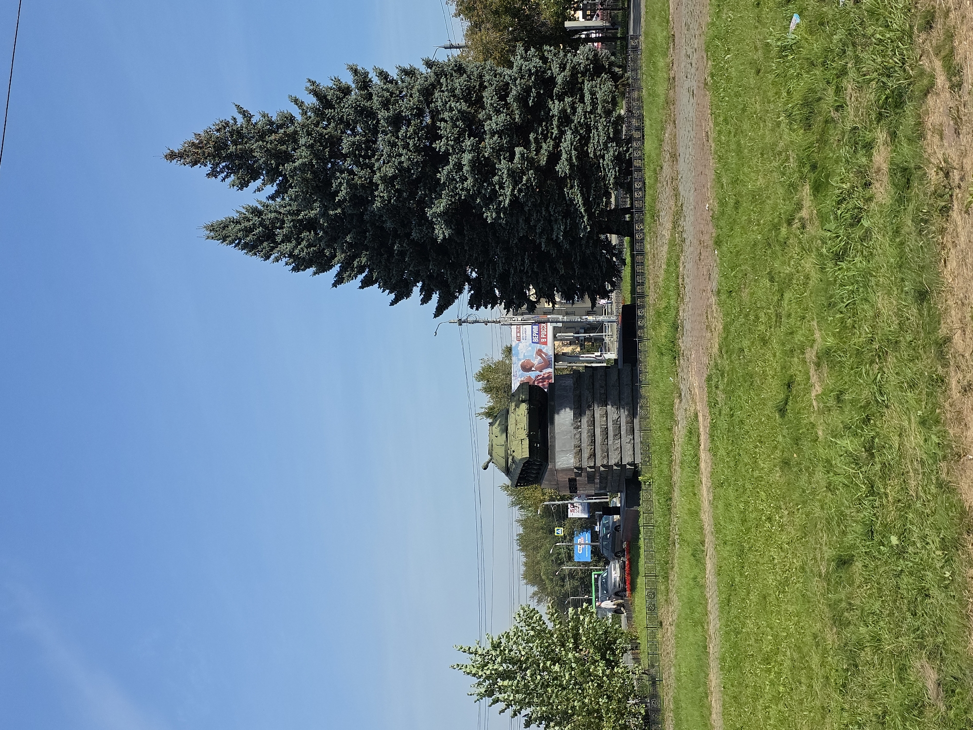 This image captures a war monument featuring a green tank placed atop a stone pedestal. The monument is situated in a park-like setting with a grassy lawn in the foreground and mature evergreen trees to the right, likely in a public space in Chelyabinsk, Russia, given the presence of a billboard in Russian. A street with passing cars is visible behind the monument, indicating an urban environment. The sky is clear and blue.