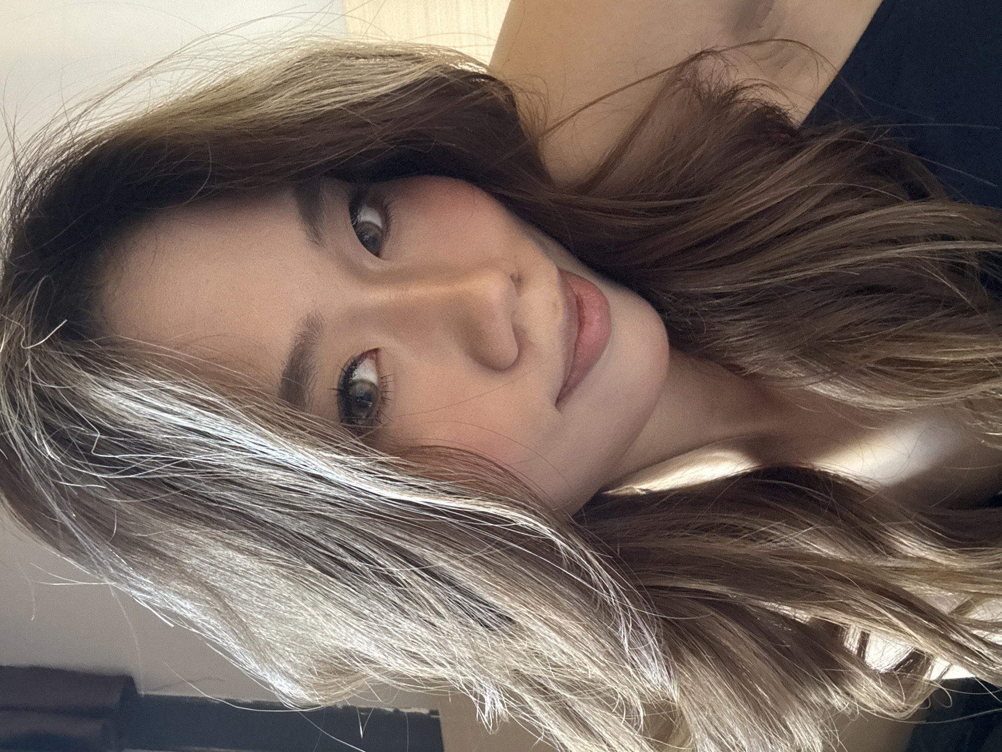 The image features a close-up portrait of a young East Asian woman, positioned diagonally across the frame. Her head is tilted upwards and to the right from the viewer's perspective, suggesting she is lying down and looking directly into the camera. She displays a soft, engaging expression, with her full lips slightly parted in a gentle smile.
Her large, dark, almond-shaped eyes are a focal point, gazing intently at the viewer, with visible eyelashes that appear enhanced with mascara. Her nose is small and well-defined, and her eyebrows are dark and neatly groomed. Her skin appears smooth and clear, with a light complexion and a touch of rosy blush on her cheeks, indicating natural-looking makeup.
Her long, wavy hair is a prominent feature, styled with considerable volume and cascading around her face and shoulders. The hair is a multi-toned brown, interspersed with distinct lighter, almost ash-blonde or silvery-blonde highlights, particularly around the front and sides, creating depth and texture. Fine, wispy hairs are visible against her skin and hair.
A small section of a dark, possibly black, garment is visible on her upper body. The background is largely indistinct and light-colored, suggesting an indoor setting with soft, diffused lighting that highlights her features without harsh shadows. The overall mood is calm and intimate. No text is visible in the image. While the context of Yuzhong District, China, was provided, there are no specific visual cues within the image to confirm this location.