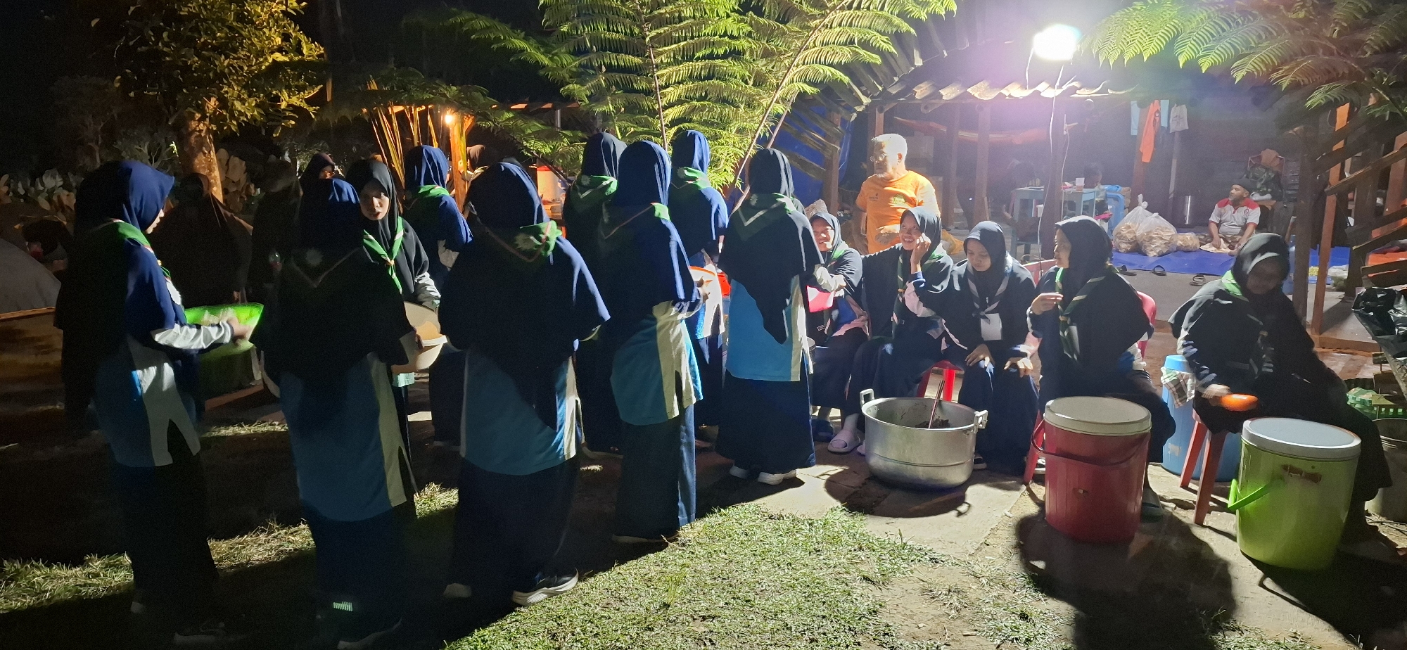 In Karang, Indonesia, a group of young individuals, likely scouts given their matching uniforms featuring blue and green elements, are gathered outdoors at night. The scene is illuminated by artificial lighting, casting long shadows on the grassy ground. Several individuals, mostly female and wearing hijabs, are seated on chairs or on the ground. Some are engaged in conversation, while others appear to be observing or participating in an activity.
In the foreground, two individuals in uniform are standing and facing away from the camera, one holding a green container. To their right, a large aluminum pot sits on the ground, possibly containing food. Several large, cylindrical containers, one red and one green, are positioned around the pot, suggesting a meal preparation or distribution setup. In the background, a man in an orange shirt is visible standing near a simple wooden structure, possibly a shelter or stall. There are also stacks of what appear to be snacks or food items on a blue tarp in the mid-ground. The environment appears to be a rural or semi-rural area, with trees and foliage visible, and the overall atmosphere suggests a communal gathering or event taking place after dark.