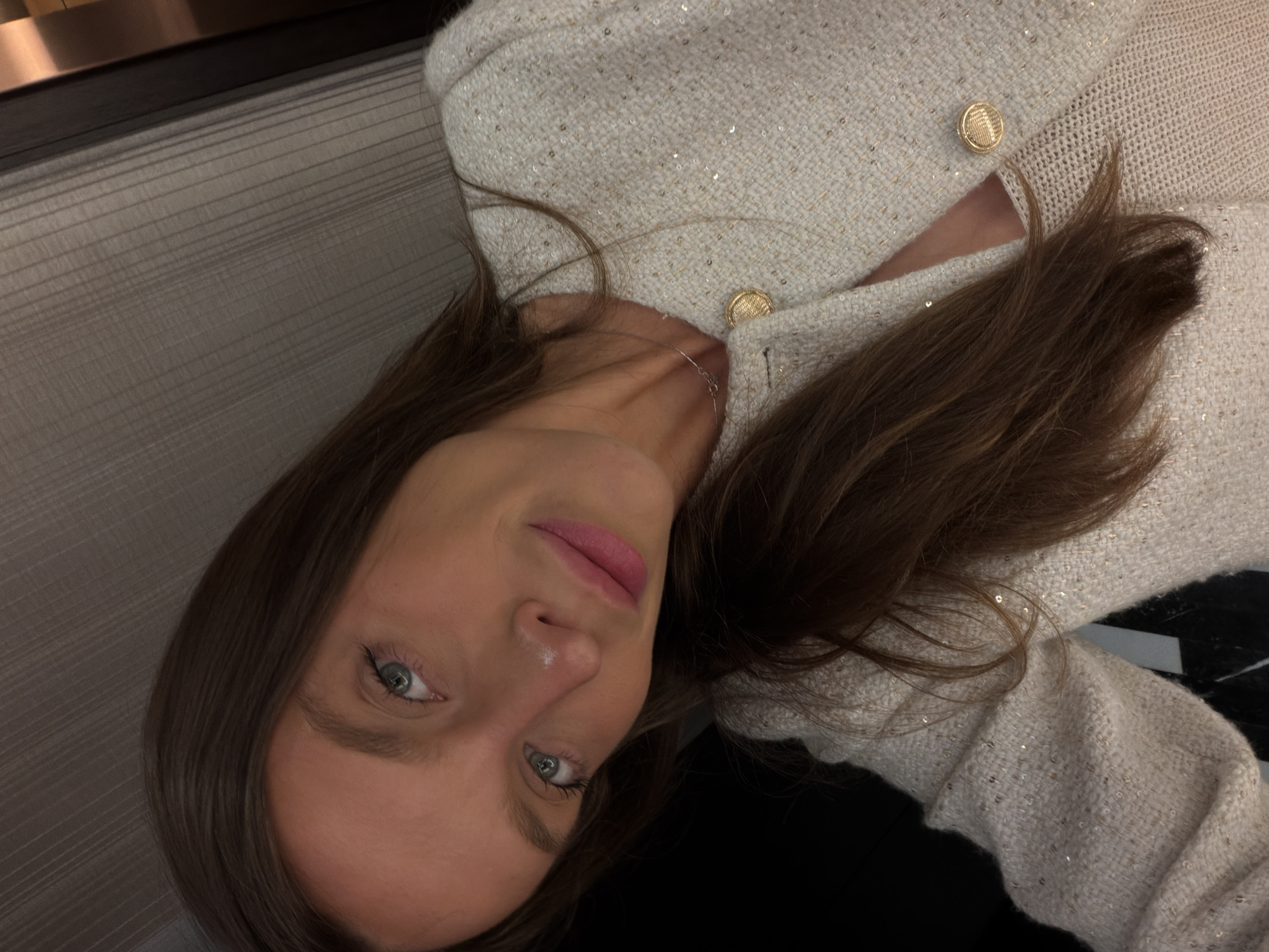 The image depicts a close-up, angled selfie of a young woman with long, dark brown hair and light blue eyes. Her lips are pink and slightly parted, and a small white mark is visible on the bridge of her nose. She is wearing a cream or off-white textured jacket, possibly tweed, which is adorned with numerous small, shimmering sequins or metallic threads. The jacket has at least two prominent gold-colored, circular, textured buttons on its front closure. A thin, delicate silver necklace is visible around her neck. The background on the upper right side features a light grey-beige wall with a subtle vertical striped texture, adjacent to a dark vertical element. The lower left and central background areas are dark and indistinct, suggesting an indoor setting. The lighting appears to be artificial, illuminating her face from the front. The provided information indicates the location as the city of Budva, Montenegro, though no specific visual cues within the image confirm this geographical detail.