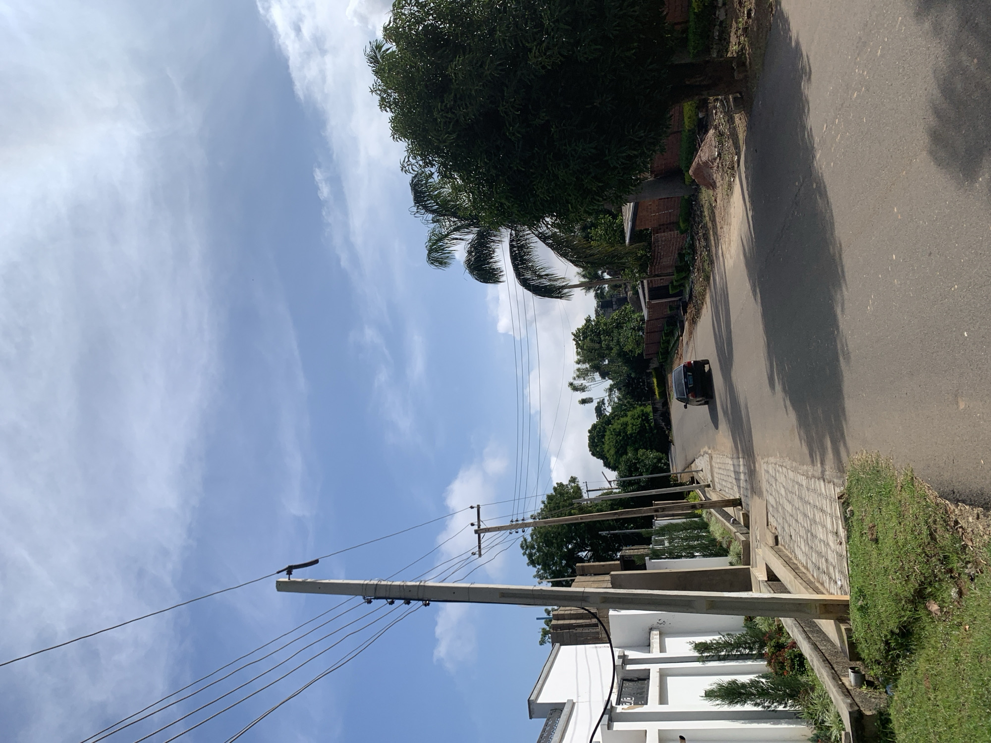 The image depicts a street scene in Jos, Nigeria, under a bright, cloudy sky. A paved road curves into the distance, bordered by a sidewalk and a grassy verge on the left. On the left side of the road, a white building with a distinct architectural style is visible, featuring a decorative white wall and manicured hedges. Utility poles with multiple overhead wires are prominent features along the street, with one particularly large concrete pole in the foreground. A dark-colored sedan is seen driving away from the camera on the road. Further down the street, a brick wall with a fence and various trees, including palm trees, are present. The overall environment suggests a residential or semi-residential area within the city.