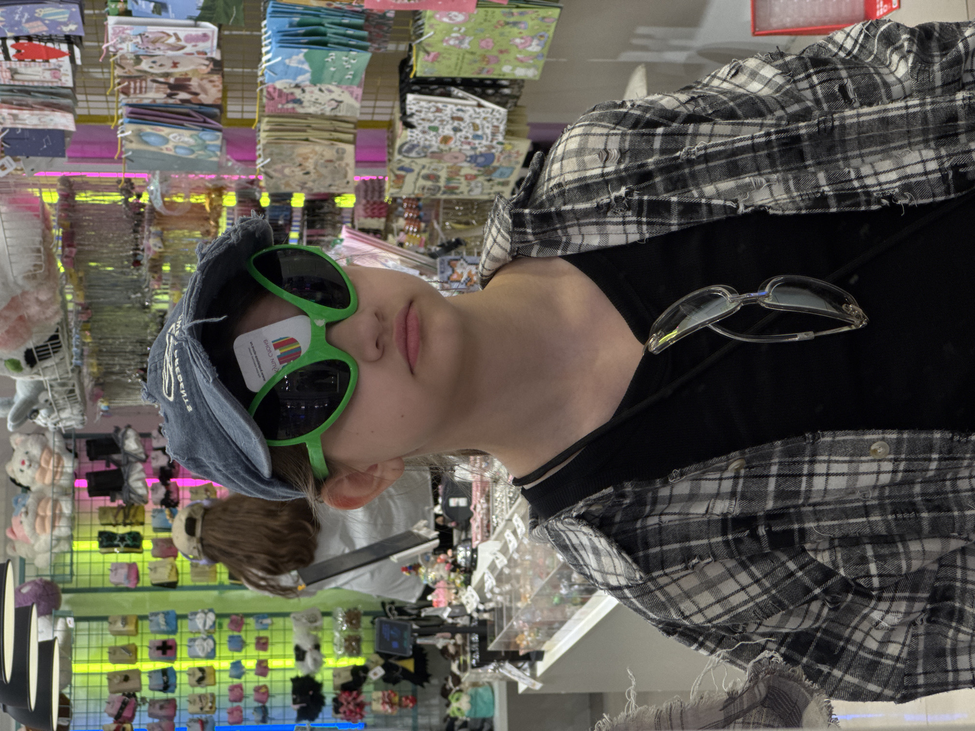 A young person wearing a denim baseball cap and bright green sunglasses is in a store, likely in Moscow, Russia given the type of merchandise. The individual is also wearing a black t-shirt and a plaid shirt. The store sells a variety of items, including bags and hair accessories, which are visible in the background. The lighting in the store is bright and somewhat fluorescent. The picture appears to be taken at a slightly tilted angle.