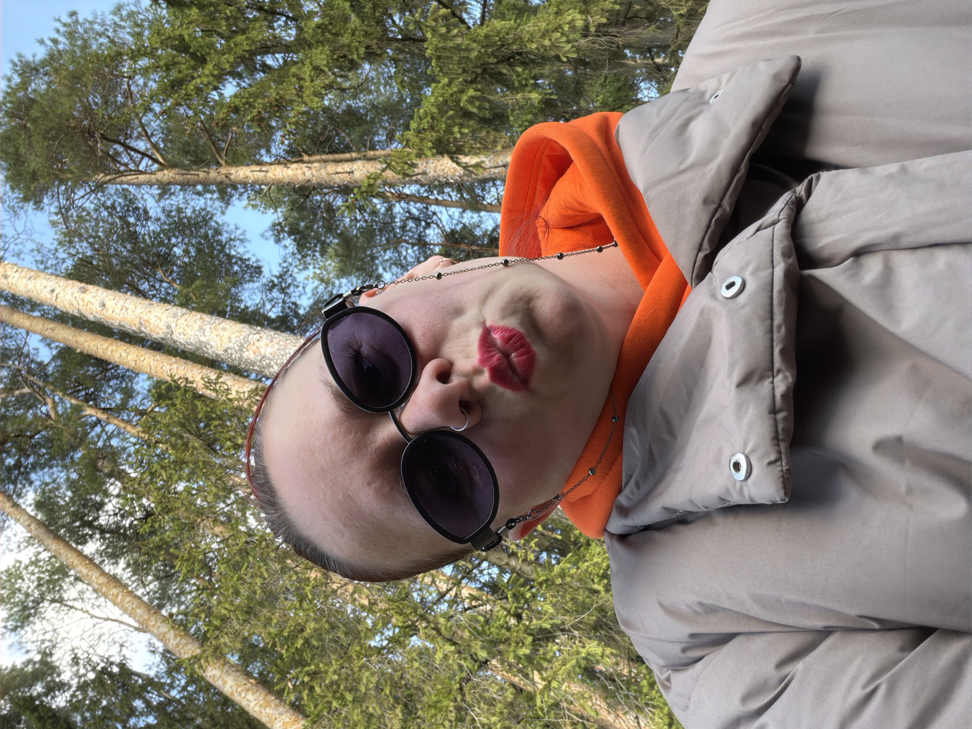 A person wearing sunglasses, an orange hoodie, and a grey puffer jacket is in an outdoor setting. Tall trees with green foliage are visible in the background, with patches of blue sky. The photograph appears to be taken from a low angle, looking up at the person and the trees. The person has bright red lipstick and is pouting slightly. The exact location within Chaikovsky, Russia is not discernible from the image.